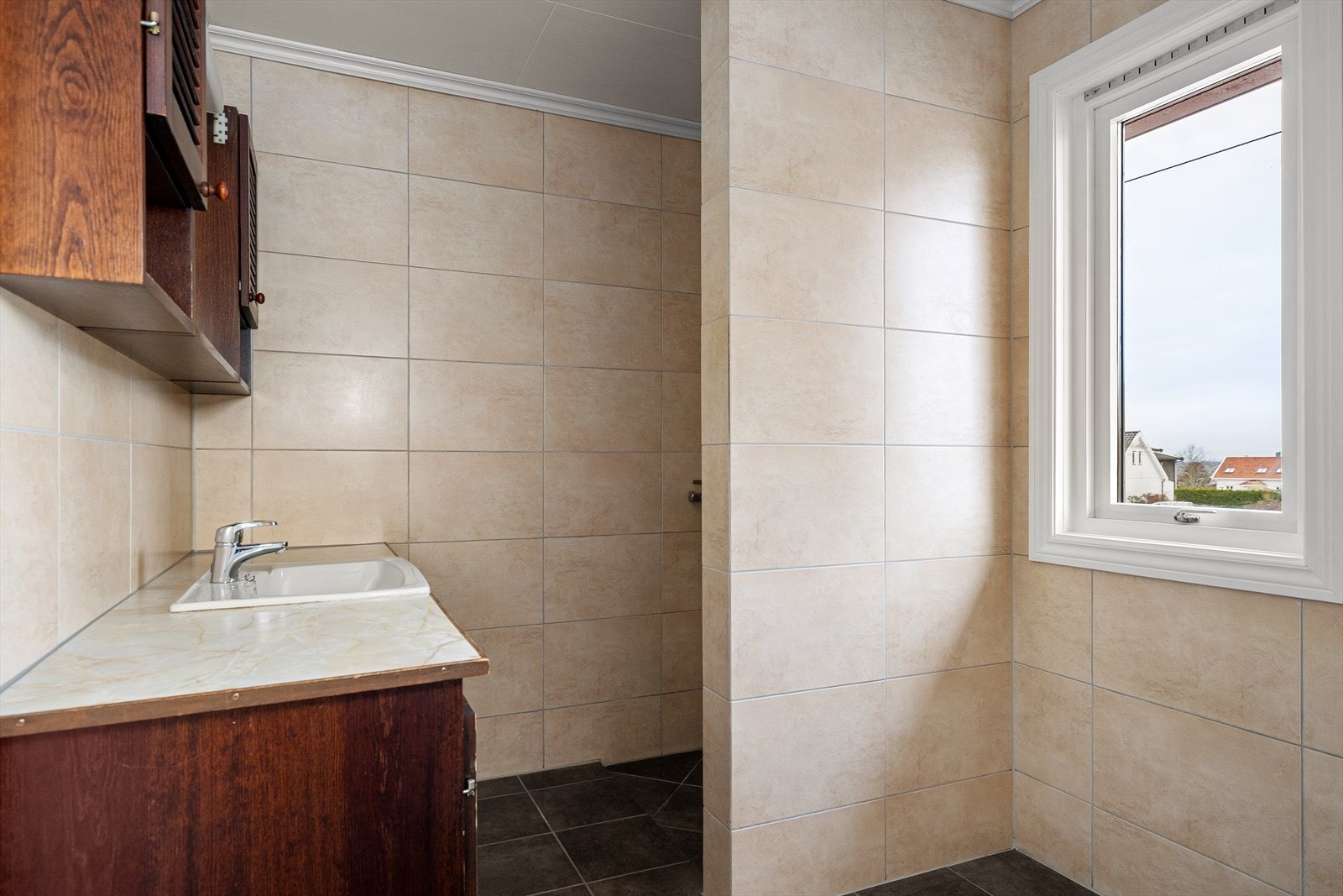 Romslig baderom med flislagte vegger fra 2006 i lyse, varme toner og naturlig lys fra stort vindu. Bak skilleveggen ligger dusjsonen diskret plassert. Galleribilde