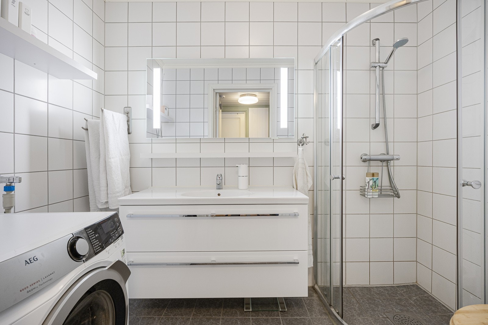 Leiligheten har et velholdt og pent bad med hvite fliser på veggene, hvitmalt panel i himlingen og grå fliser på gulv med gulvvarme. Galleribilde