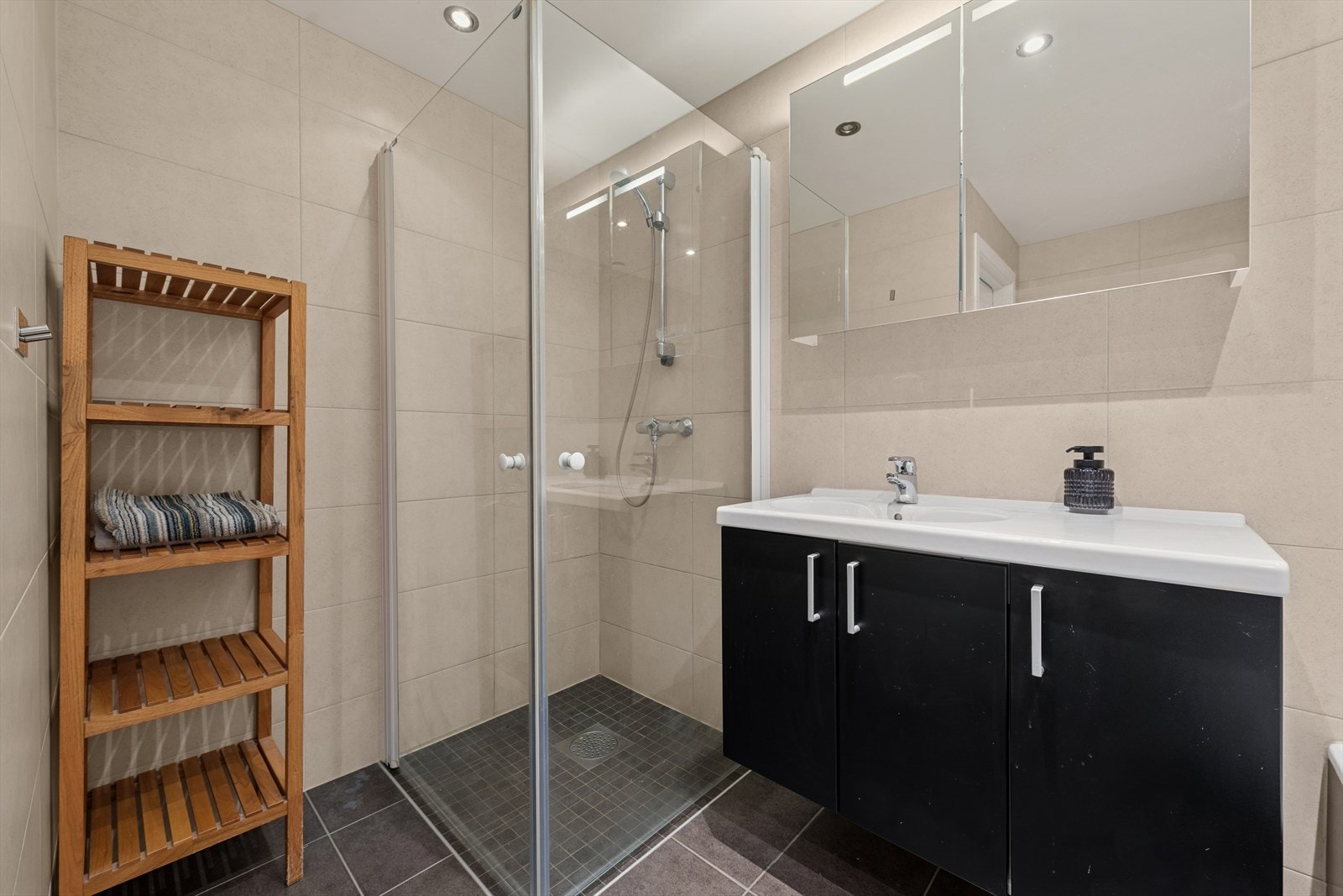 Rommet er utstyrt med dusjhjørne med svingbare skjermvegger, innmurt flissatt badekar med dusj, vegghengt klosett, hel nedfelt servant og baderomsinnredning med servantskap og speilskap med integrert belysning. Galleribilde