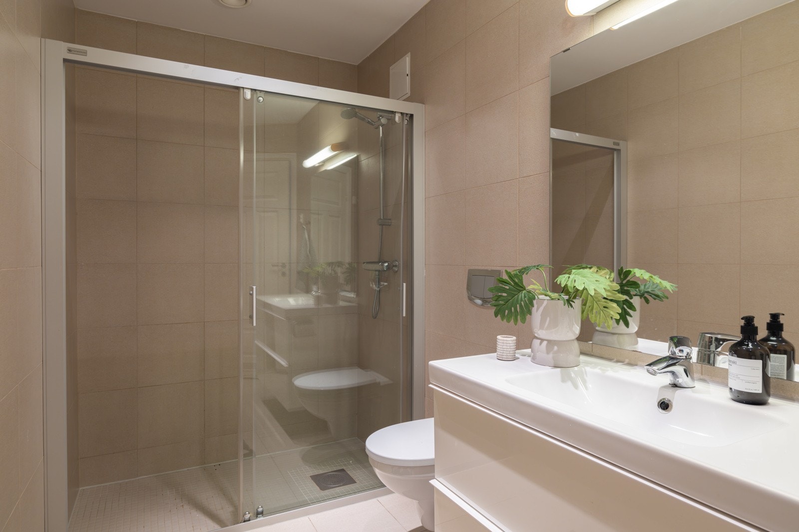 Moderne baderomsinnredning med skuffer og heldekkende servant, speil med belysning over. Baderommet er også utstyrt med romslig dusjnisje med skyvedør av glass, vegghengt toalett og opplegg for vaskemaskin. Galleribilde