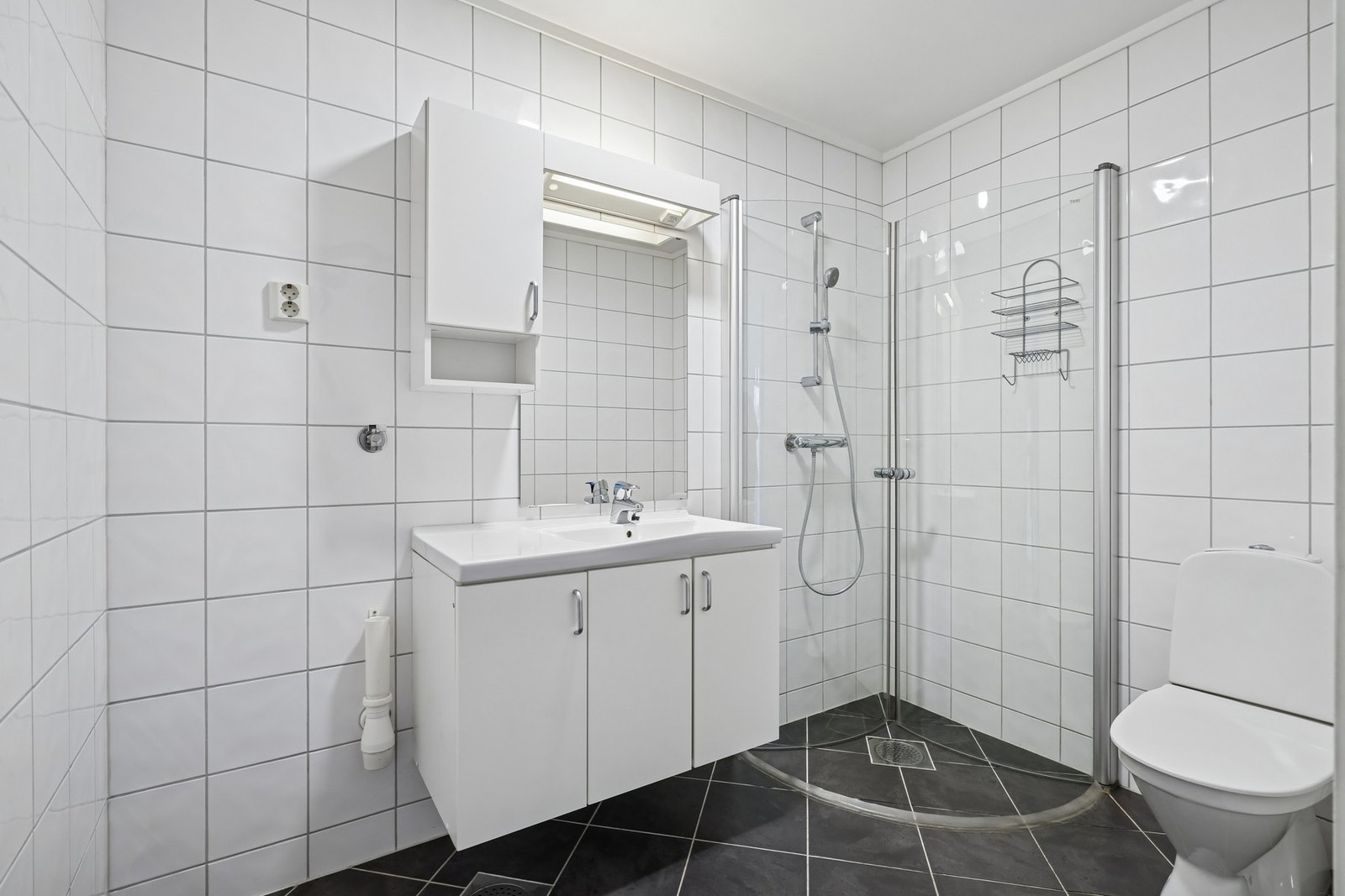 Lyst helfliset bad/vaskerom med elektriske varmekabler. Innredet med toalett, dusjhjørne, servantinnredning, et vegghengt skap og speil. Galleribilde