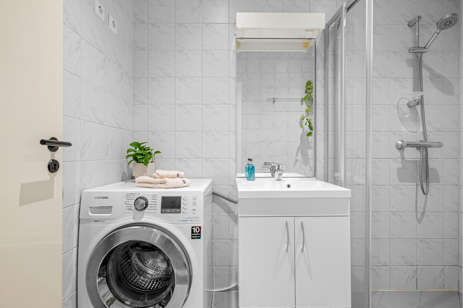 Lys baderomsinnredning med ovenpåliggende servant, speil med belysning over. I hjørnet er det plass og opplegg for vaskemaskin. Galleribilde