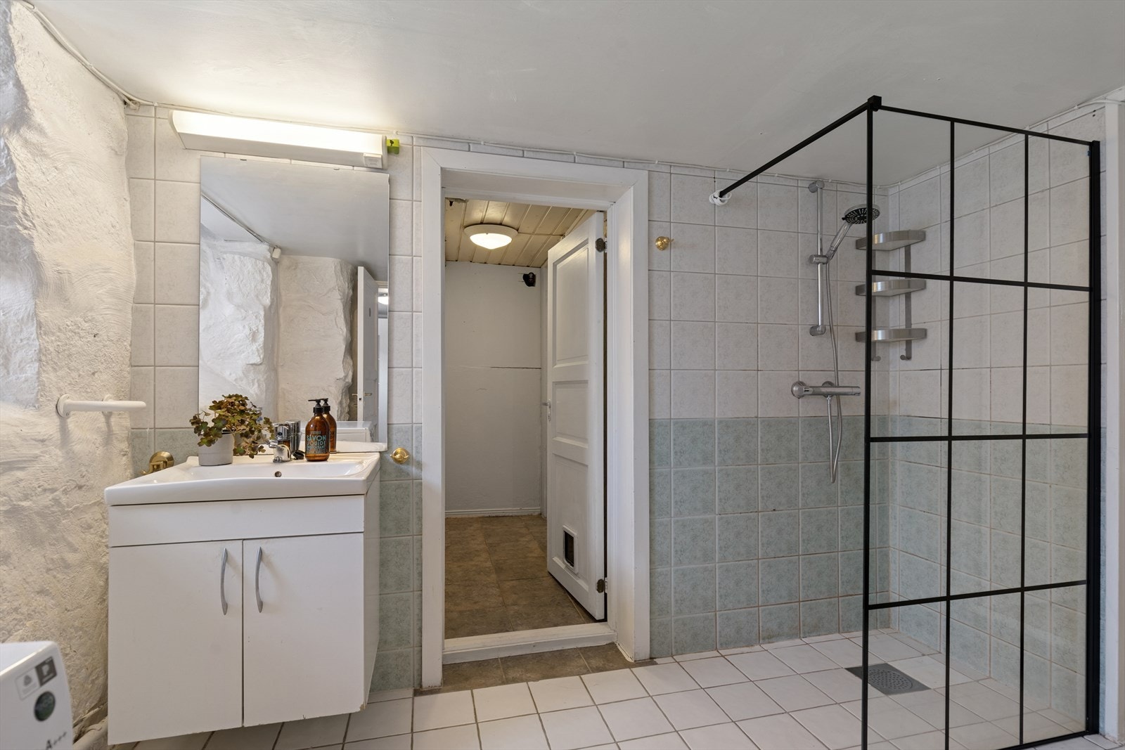 Bad i kjellerenheten med flislagt gulv, dusjhjørne og opplegg for vaskemaskin. Galleribilde