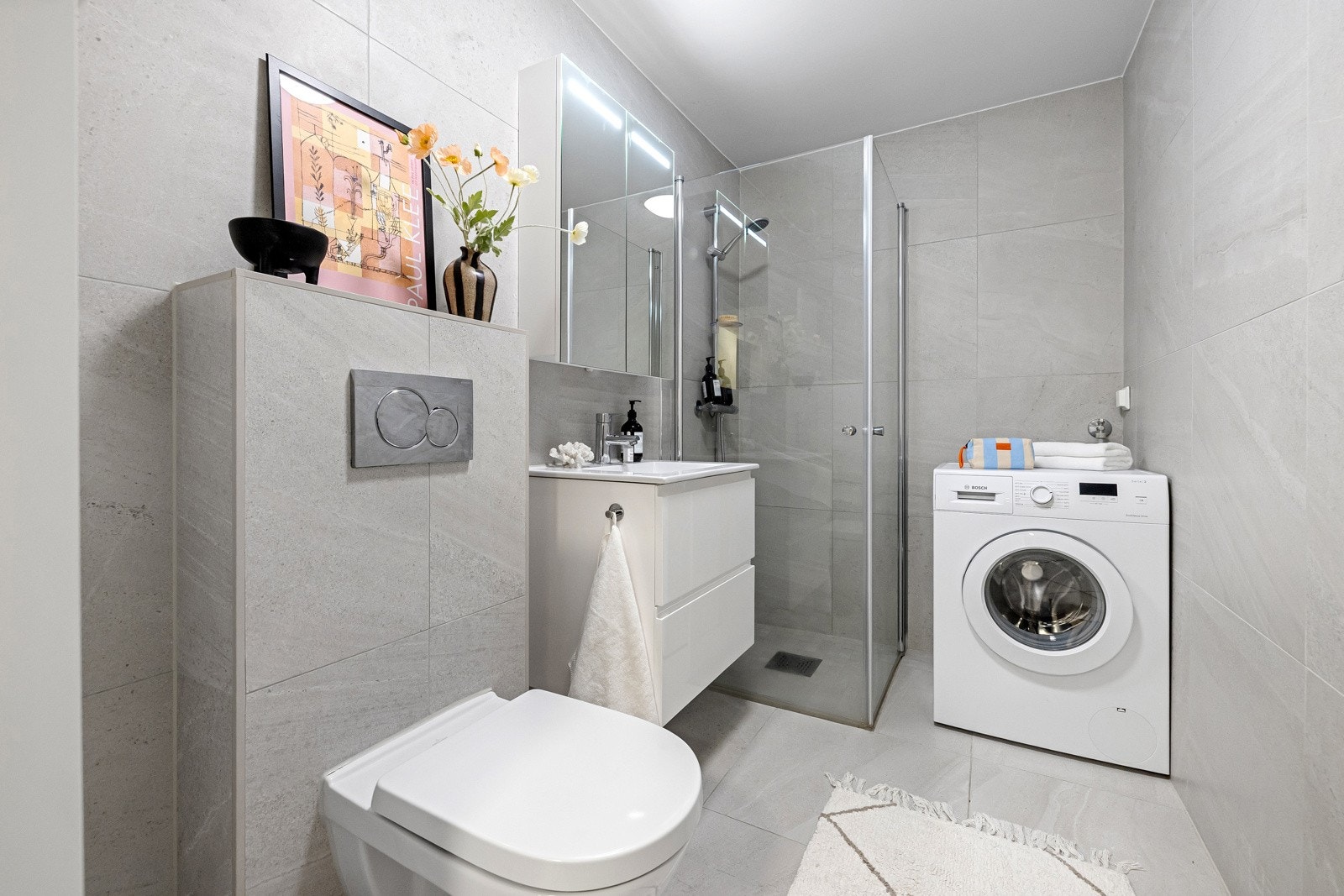 Badet er innredet med servant med underskuffer, speilskap med moderne belysning, vegghengt toalett og dusjhjørne med innfellbare dusjdører. Galleribilde