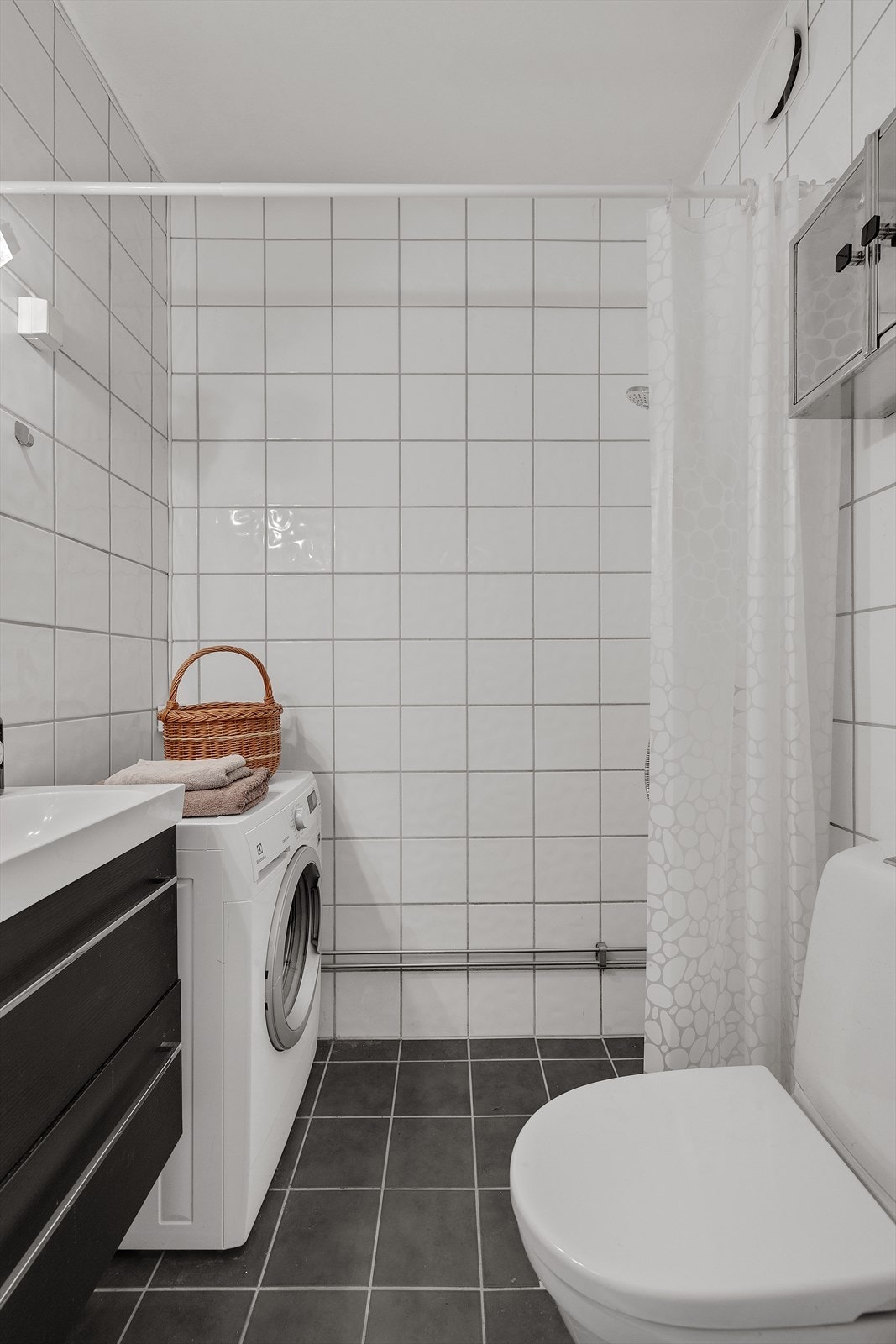 Badet inkluderer også gulvmontert klosett, dusjhjørne med forheng, og opplegg for vaskemaskin. Galleribilde