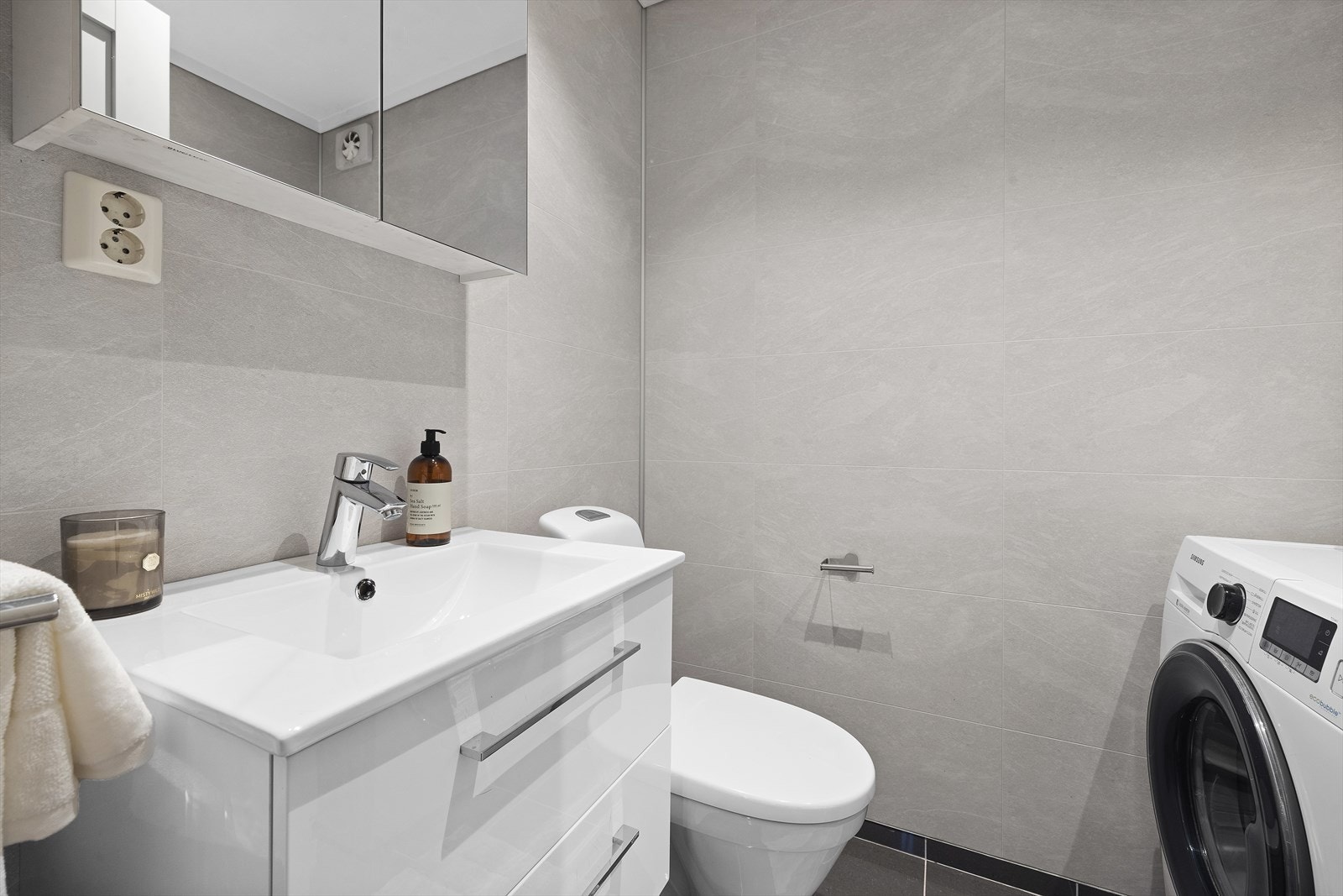 Her er det store, grå fliser på gulv og lyse baderomsplater på vegger, moderne baderomsinnredning og opplegg for vaskemaskin og tørketrommel. Galleribilde