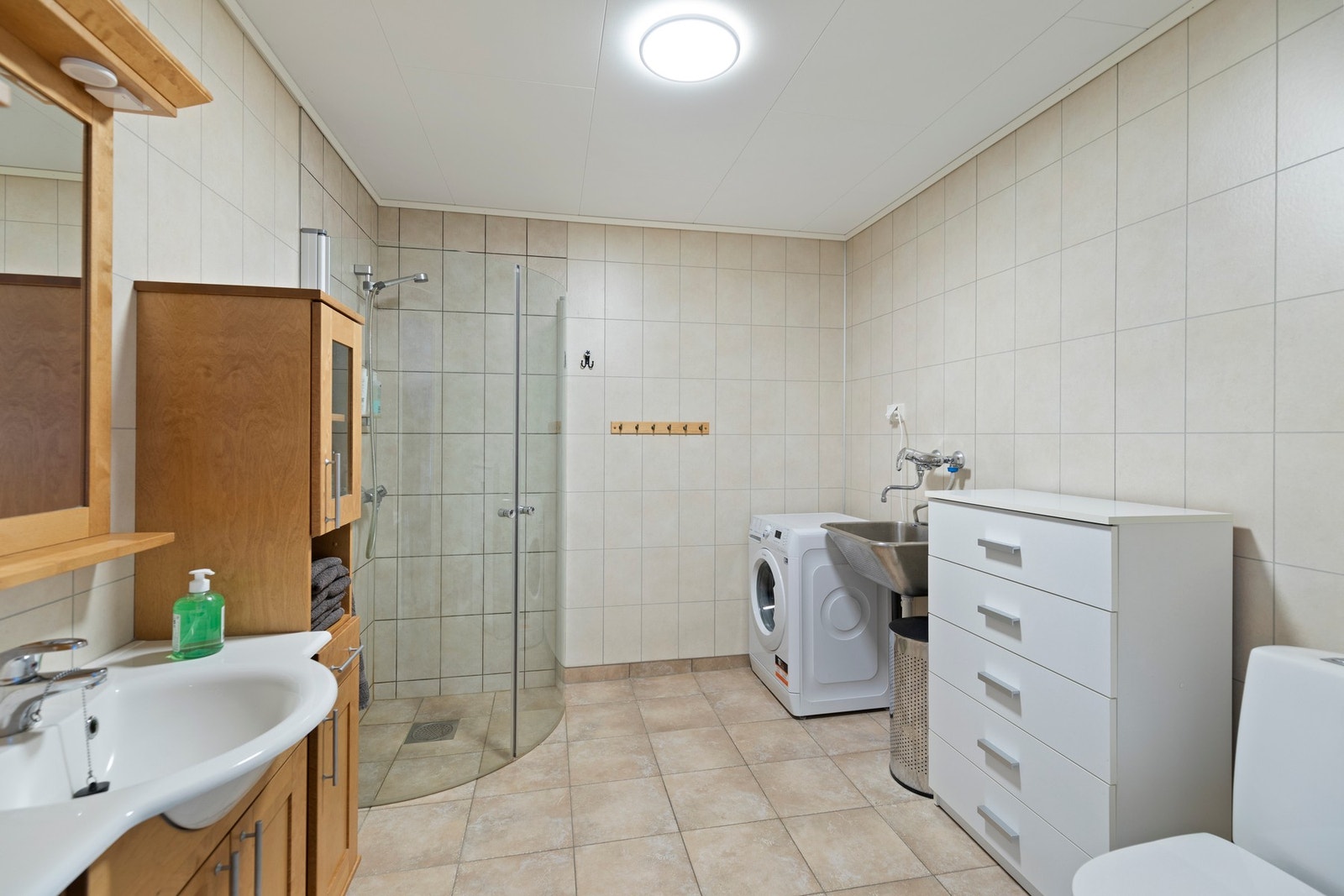 Kombinert bad og vaskerom med baderomsinnredning, dusjnisje med glassdører, wc, utslagsvask og opplegg for vaskemaskin Galleribilde