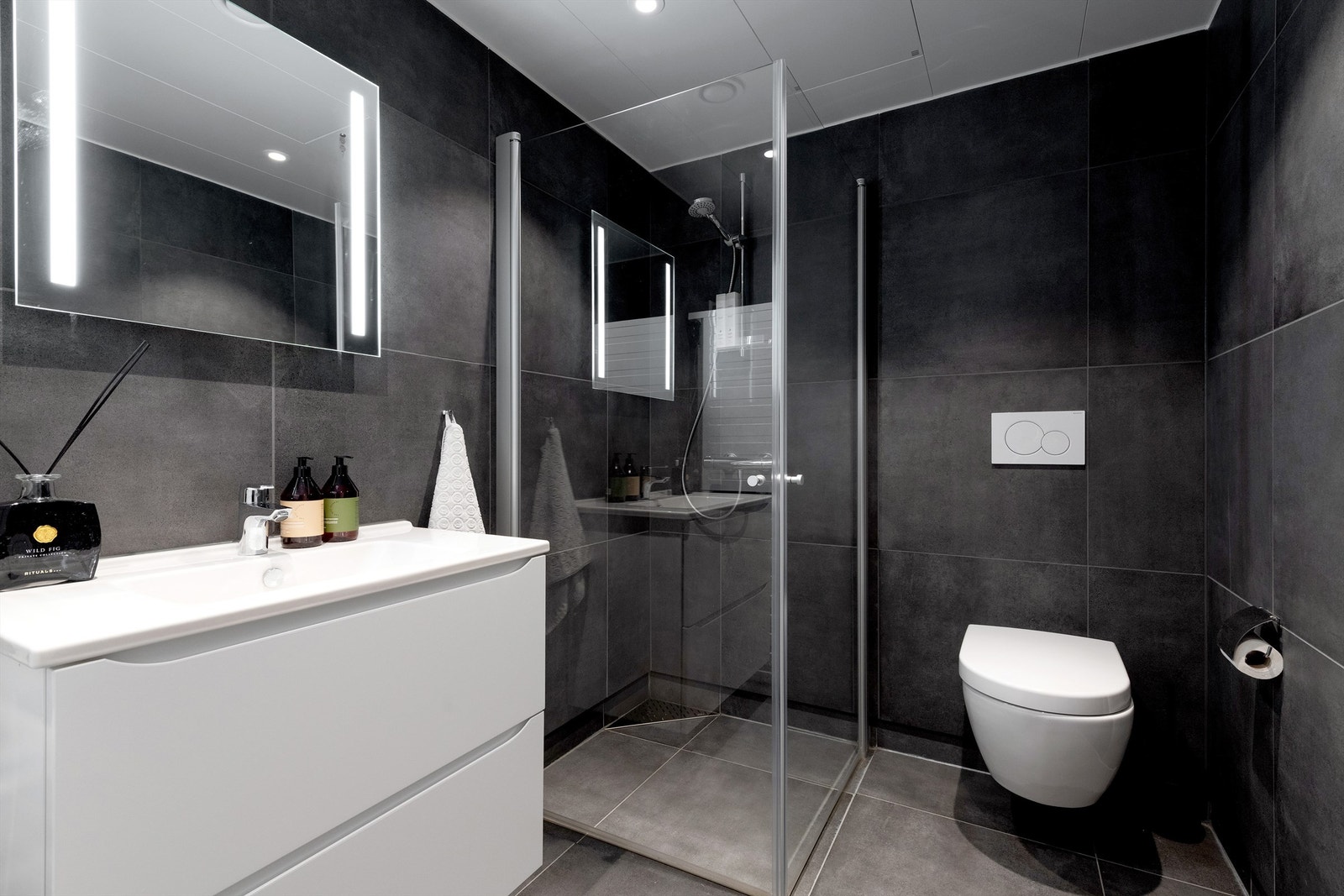 Bad med dusjhjørne, vegghengt toalett, vegghengt servantinnredning og speil med lys Galleribilde