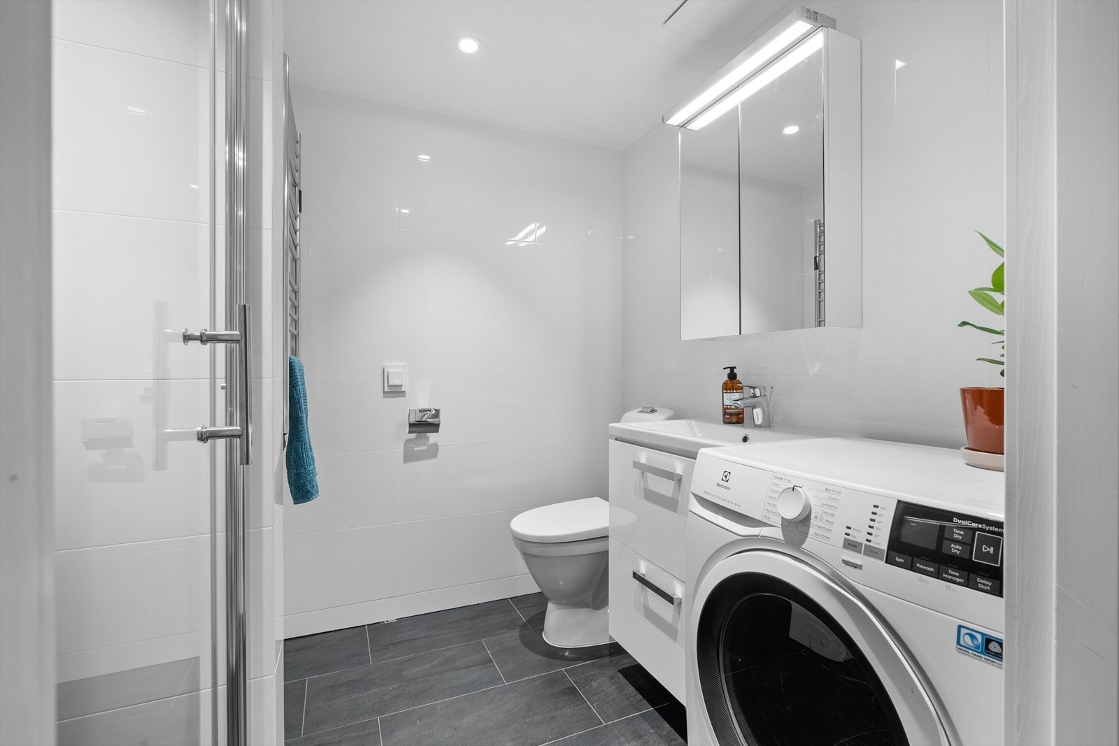 Baderomsinnredning med glatte, folierte fronter og heldekkende servant. Speilskap med lysarmatur på vegg over servanten. Det er håndkletørker montert på vegg. Galleribilde