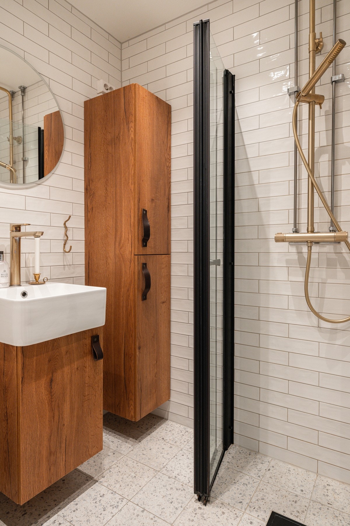 Stilrent bad med hvite veggfliser, terrazzoinspirert gulv, dusjhjørne og armatur i varm messingfinish. Galleribilde