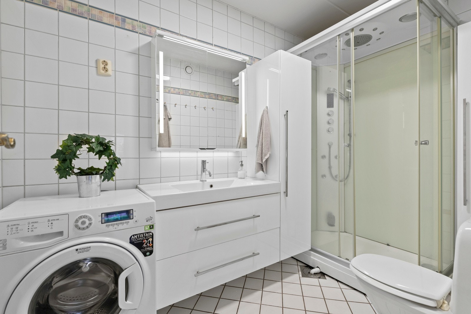 Flislagt bad som er innredet med dusjkabinett, servant i innredning, toalett og opplegg for vaskemaskin. Galleribilde