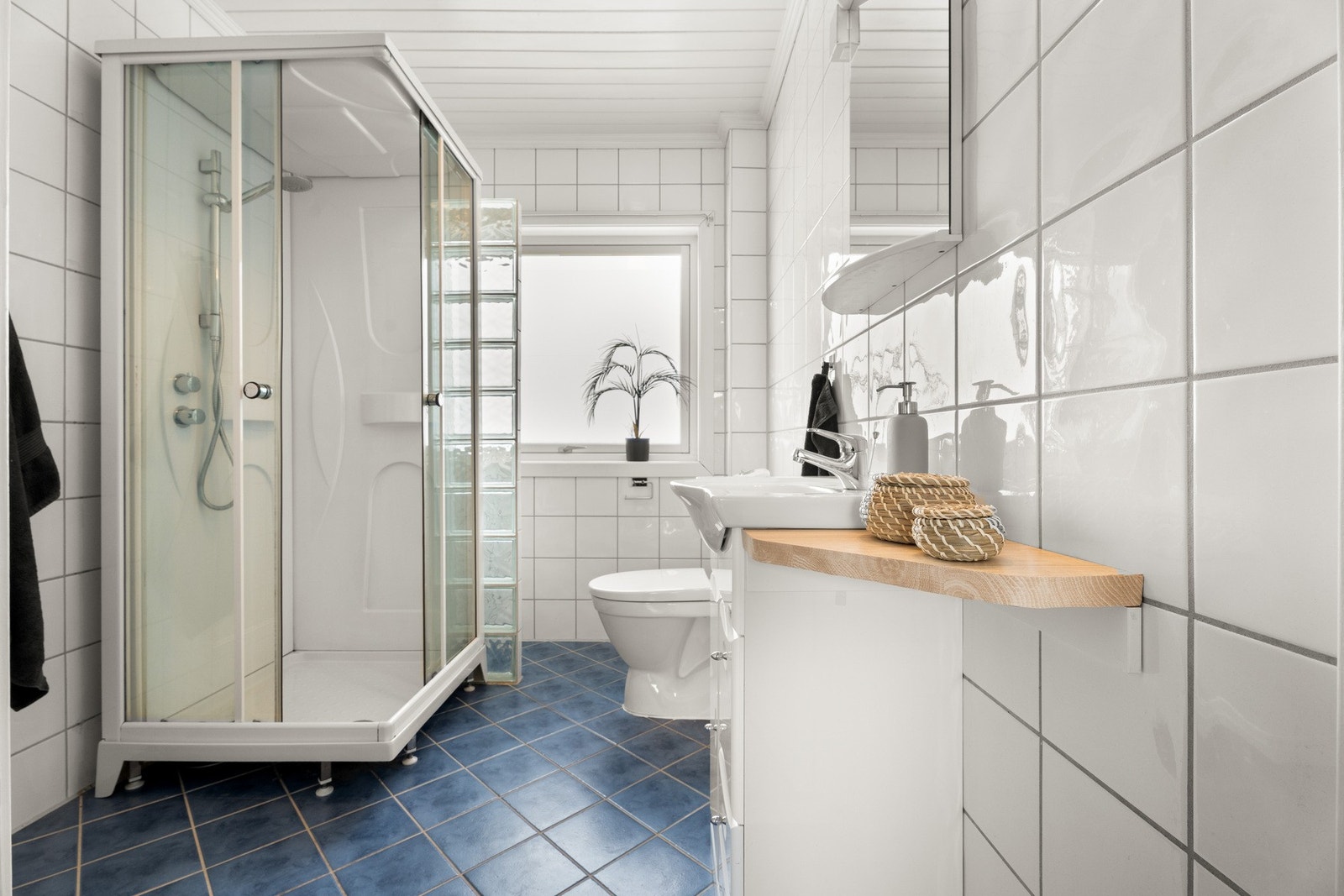 Badet fremstår som arealeffektivt og velholdt, med hvite fliser på veggene, hvitmalt panel i himlingen og blå fliser på gulv med elektrisk gulvvarme. Galleribilde