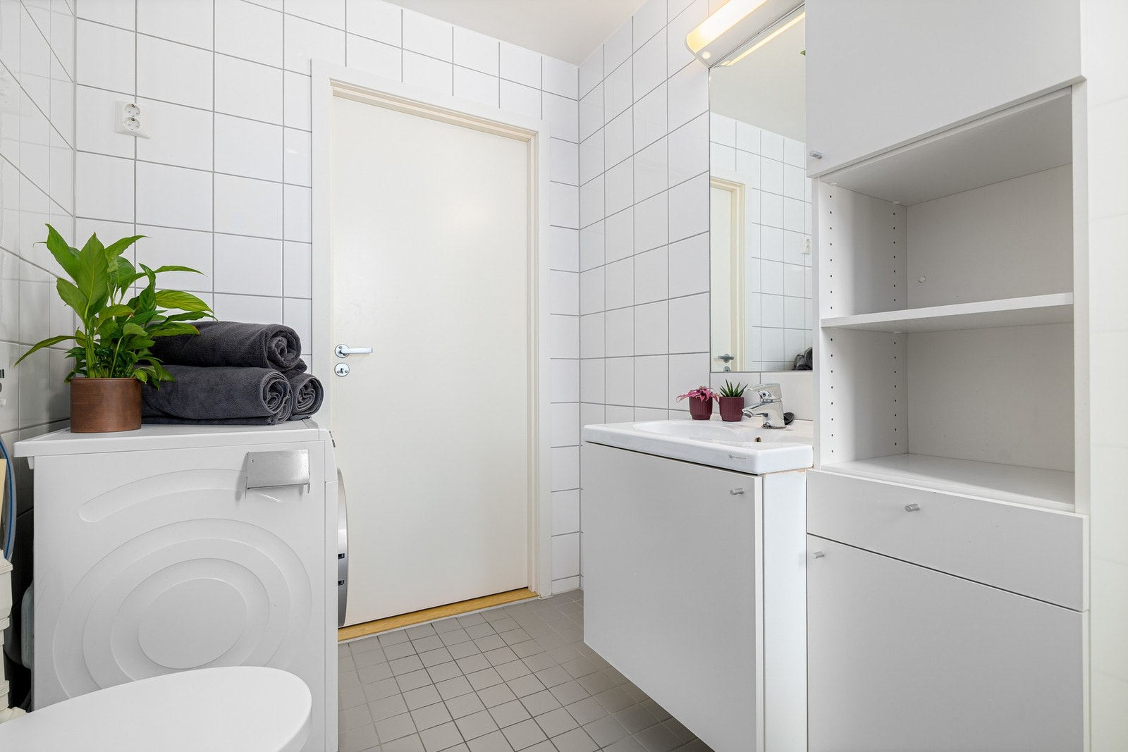 Lys baderomsinnredning med ovenpåliggende servant, speil med belysning over, samt ekstra høyskap ved siden av. Baderommet er også utstyrt med dusjhjørne med innfellbare glassdører og vegghengt toalett. Galleribilde