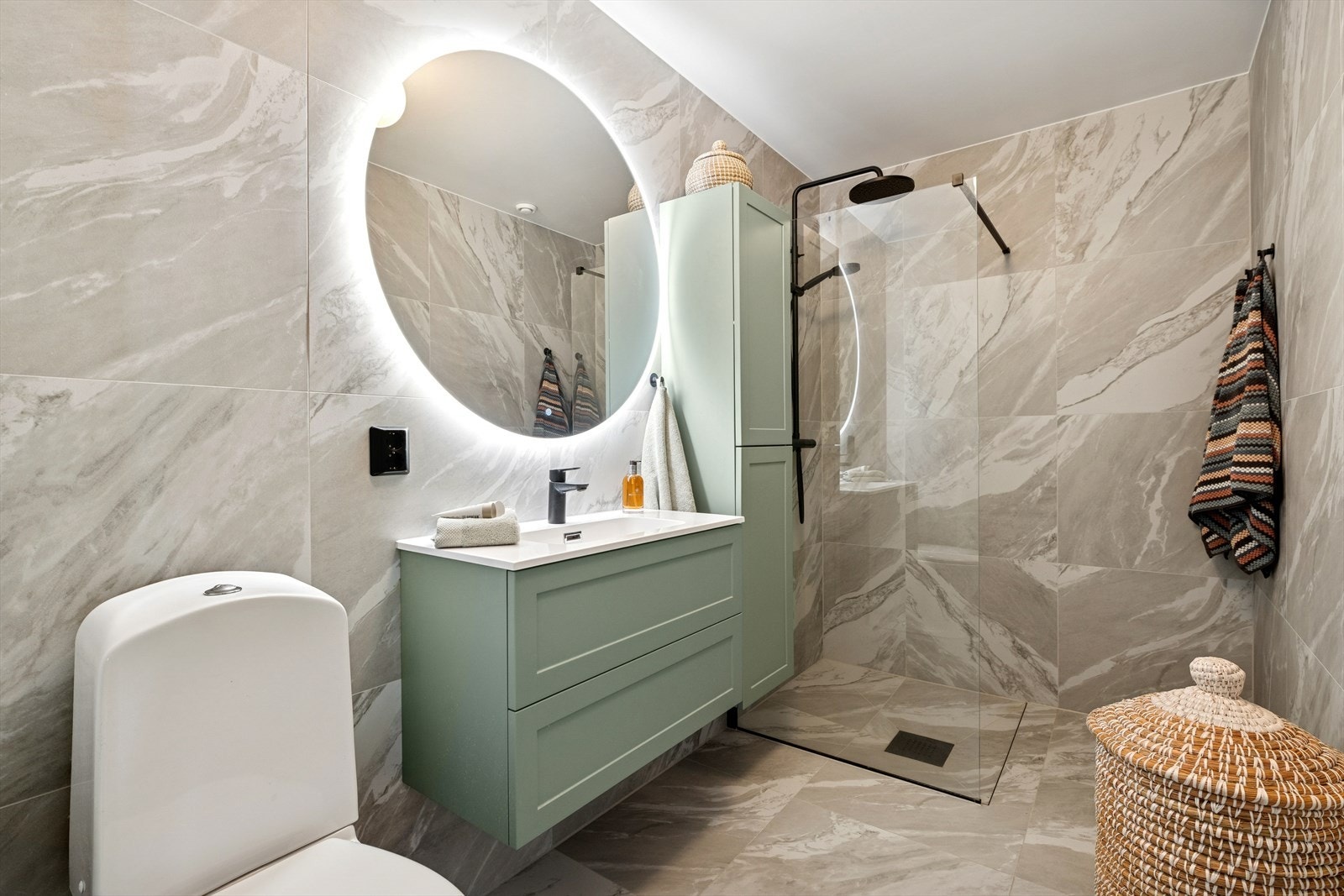 Innredningen består videre av en heldekkende servantplate på lys skuffeseksjon, veggskap i samme stil, rundt speil med belysning og toalett. Rommet har lyse og mønstrede fliser på gulv og vegger i en eksklusiv utførelse. Galleribilde