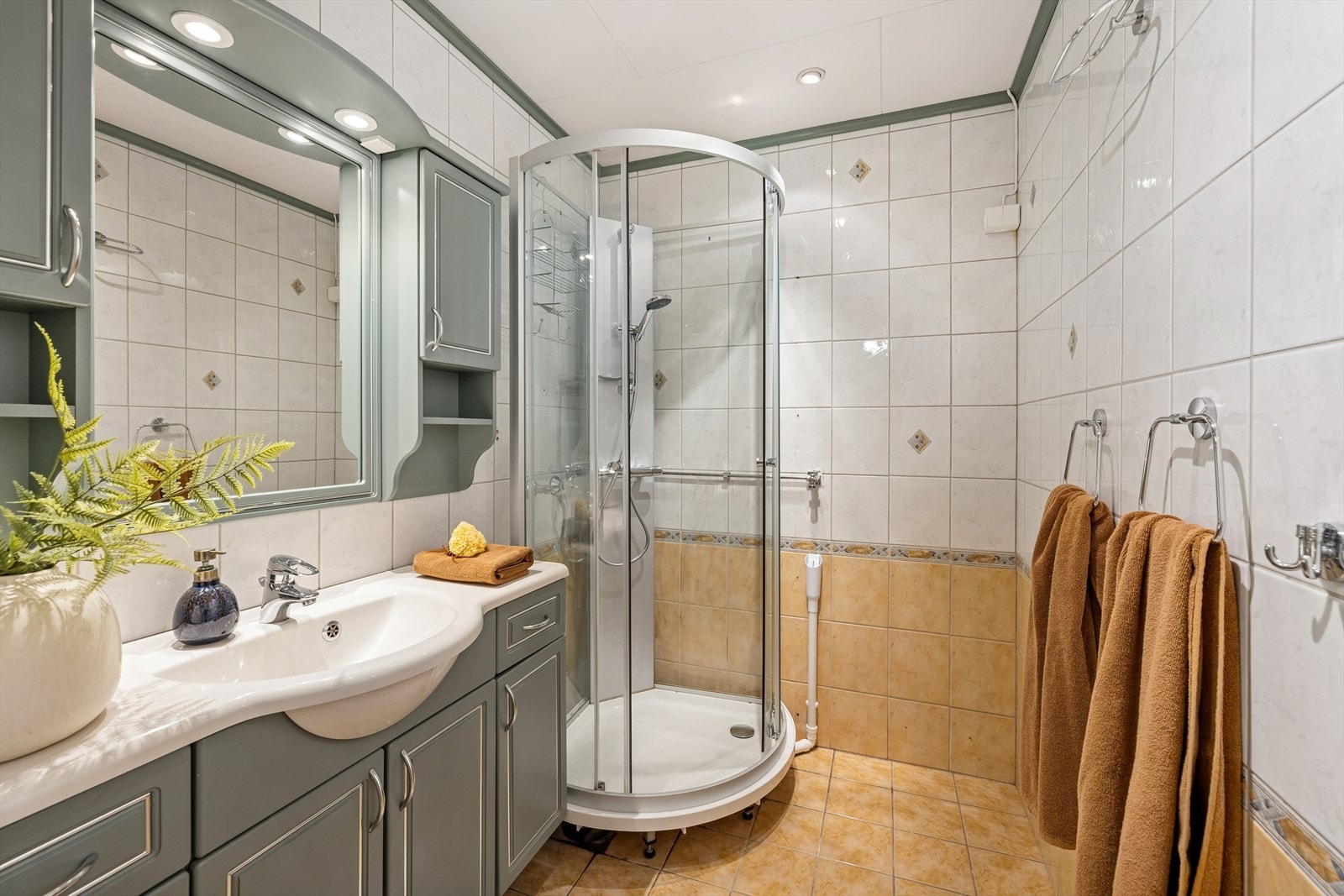 Bad med fliser på vegger og gulv med gulvvarme. Rommet er utstyrt med servant med underskap, speil med tilhørende belysning, dusjkabinett og opplegg for vaskemaskin. Det er mekanisk avtrekk og tilluft i dør. Galleribilde