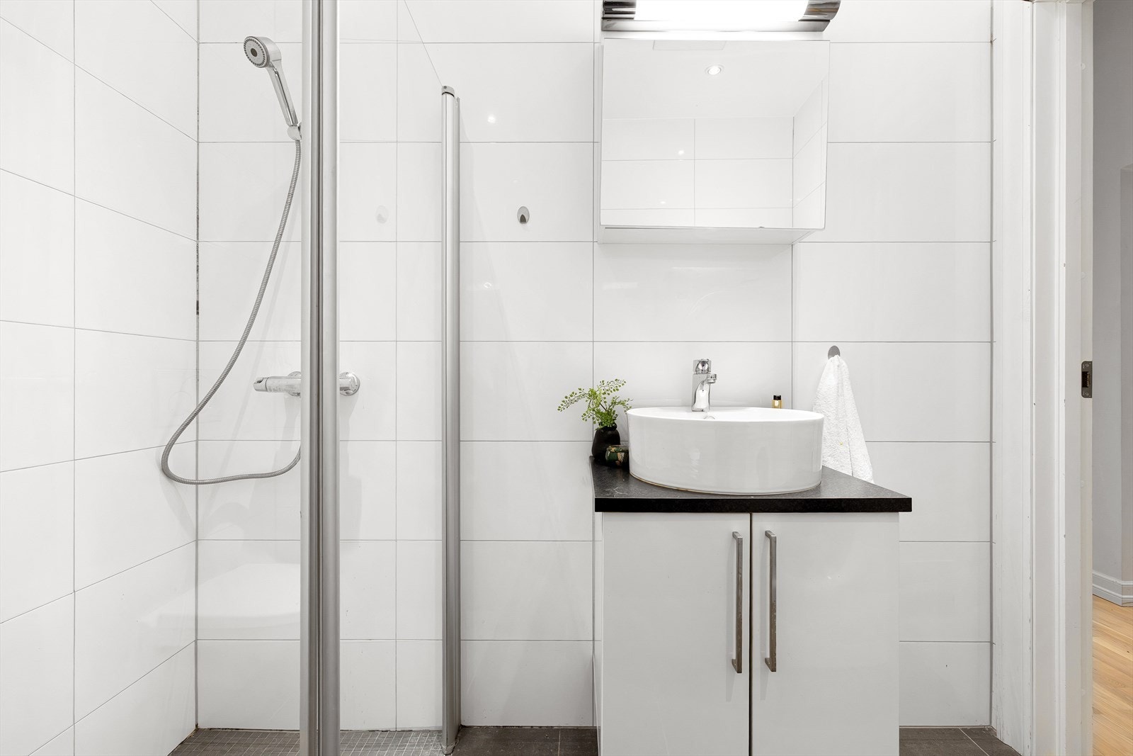 Videre er badet utstyrt med dusjhjørne med innfellbare dusjdører, veggmontert wc og opplegg for vaskemaskin. Naturlig ventilasjon via ventil og defekt elektrisk vifte. Galleribilde