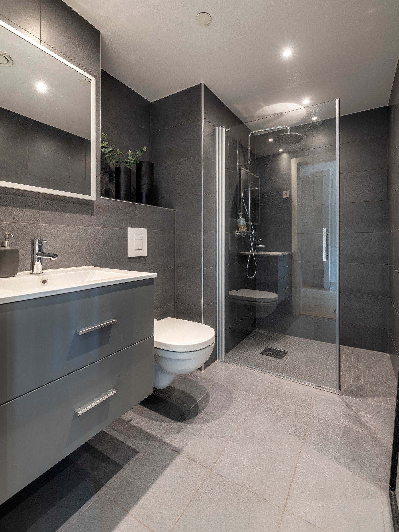 Badet har et stilrent utrykk med store vegg- og gulvfliser i grå toner, downlights, varmekabler og balansert ventilasjon. Galleribilde