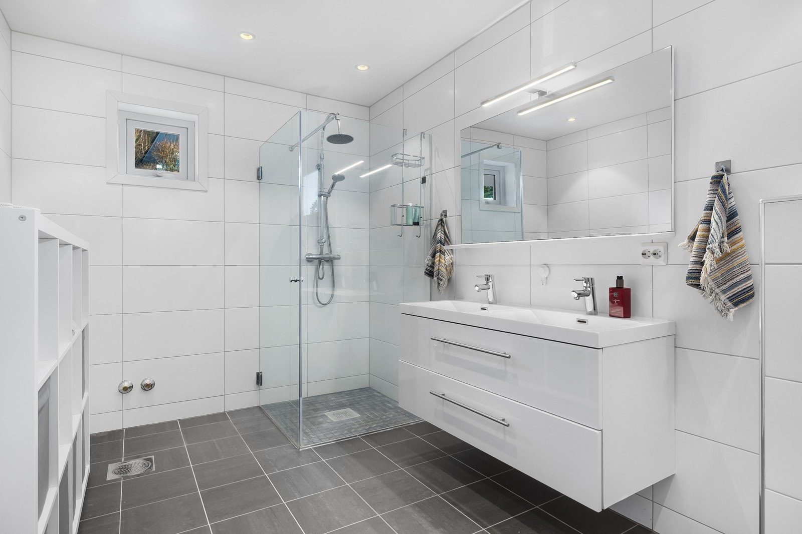 Helfliset baderom med mørke fliser på gulv og store hvite fliser på vegg. Dobbel servant med skuffer, stort speil og praktisk hjørnedusj med innfellbare glassdører. Galleribilde