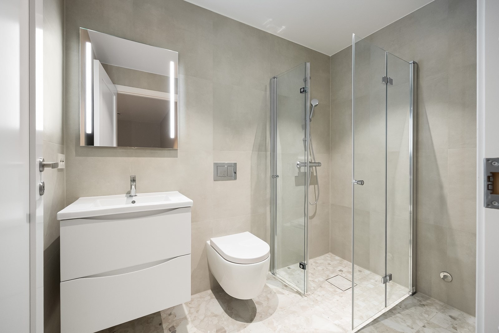 Bad med flislagte overflater, varme i gulvet, veggfestet toalett og servant samt dusjsone med dører i glass Galleribilde