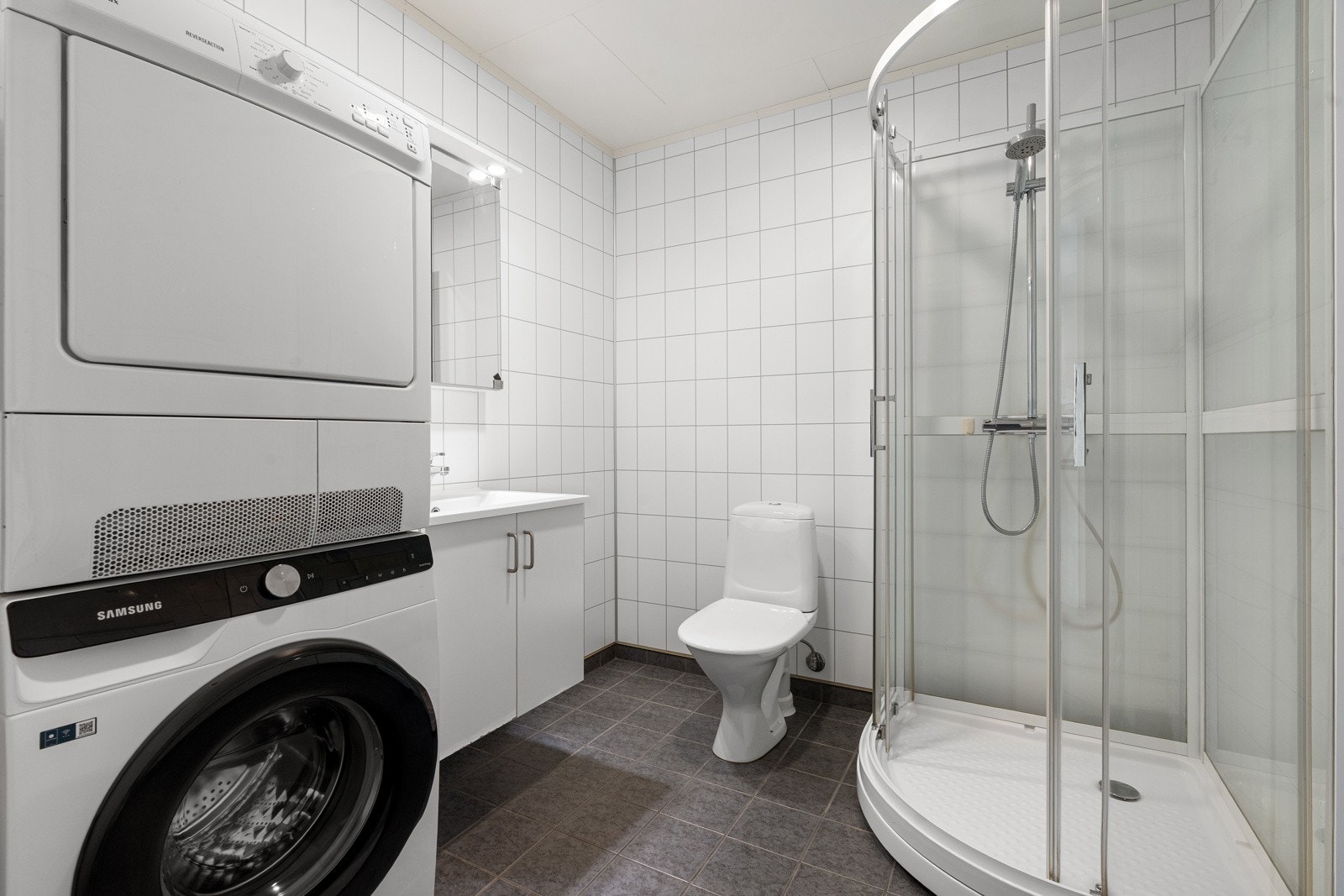 Badet er flislagt på vegger og gulv, og er innredet med vegghengt servant, dusjkabinett, toalett og opplegg for vaskemaskin.