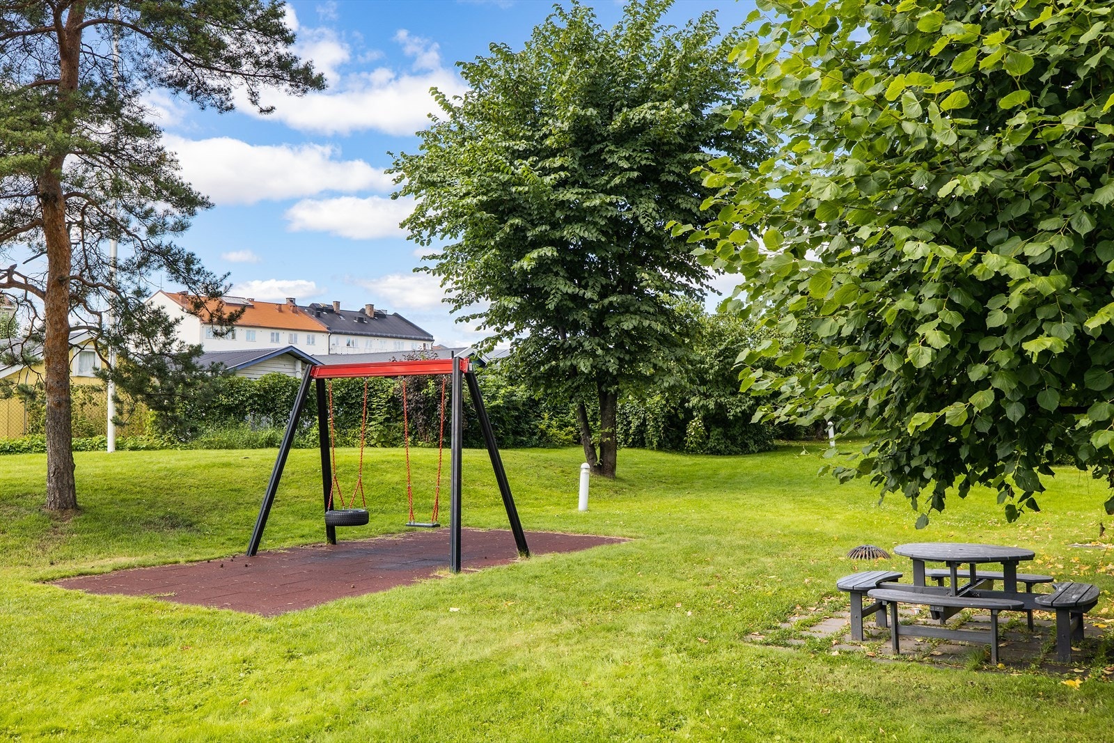 Lekeplass med huske og sitteområde ligger i et grønt parkområde. Galleribilde