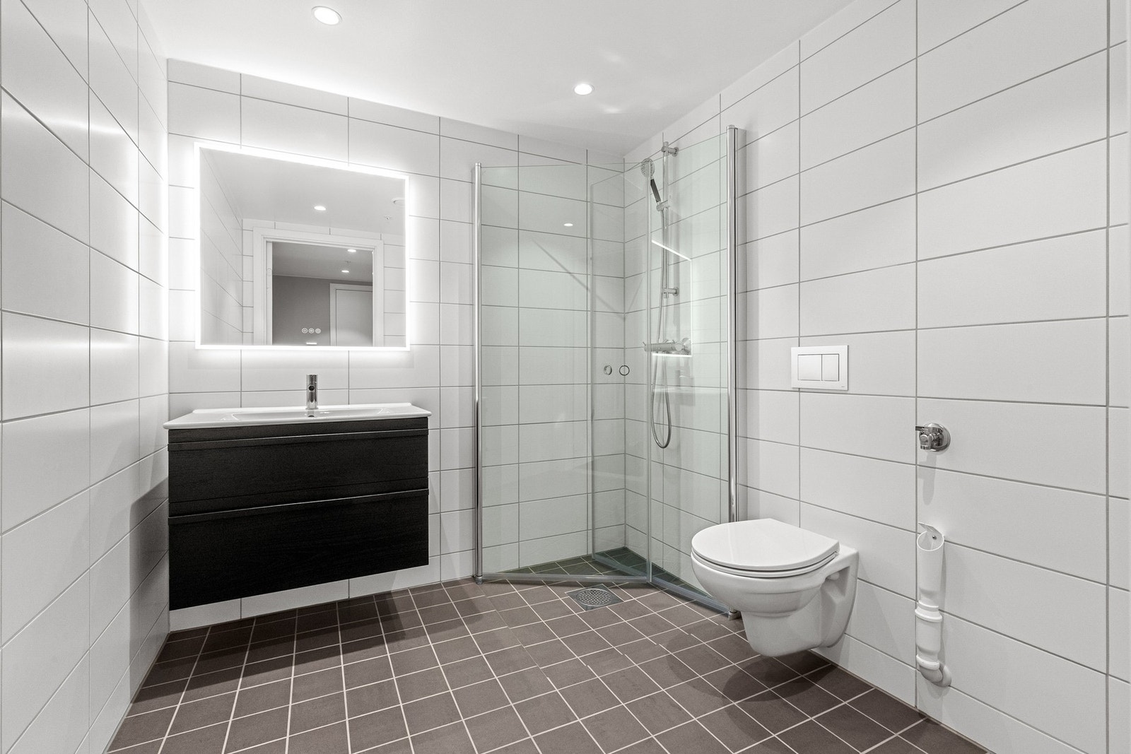 Badet ligger praktisk plassert i tilknytning til entréen. Badet er utstyrt med vegg- og gulvfliser, varmekabler og innfelt downlights. Innredningen består av servant med skuffeseksjon, speil med belysning, vegghengt toalett og dusjhjørne med innfellbare g Galleribilde