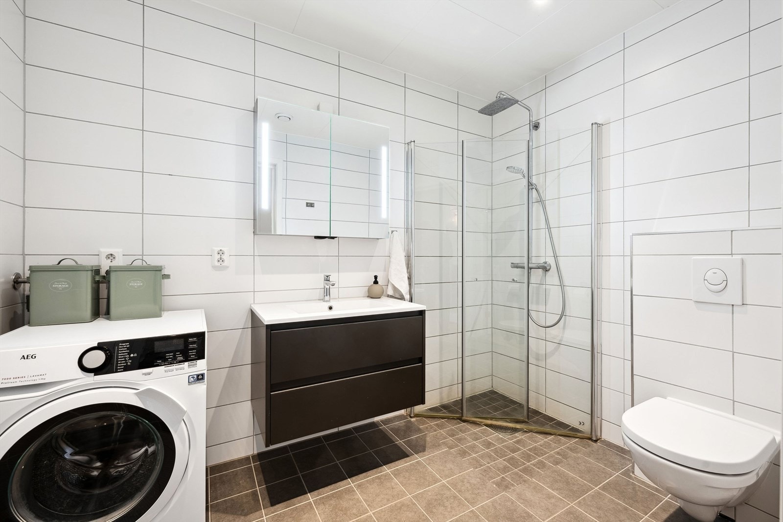 Strøkent, flislagt bad i 1.etasj. Baderomsinnredningen er sort med heldekkende nedfelt servant med rom for oppbevaring i skuffeseksjon og speil. Videre er badet innredet med vegghengt toalett og dusjnisje. Galleribilde