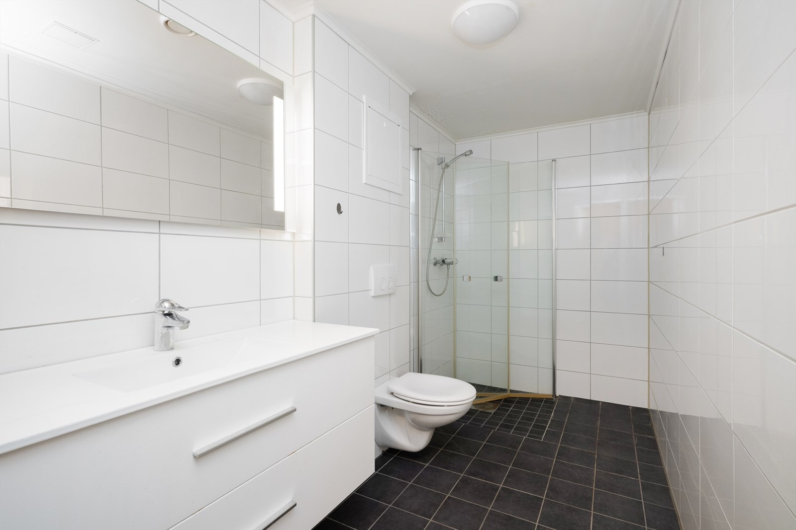 Pen og bred servantinnredning med skuffer og med nedsunket servant og speil med lys. Badet har keramisk flis på gulv og vegger, samt malte plater i tak. Avtrekk via balansert boligventilasjon. Galleribilde