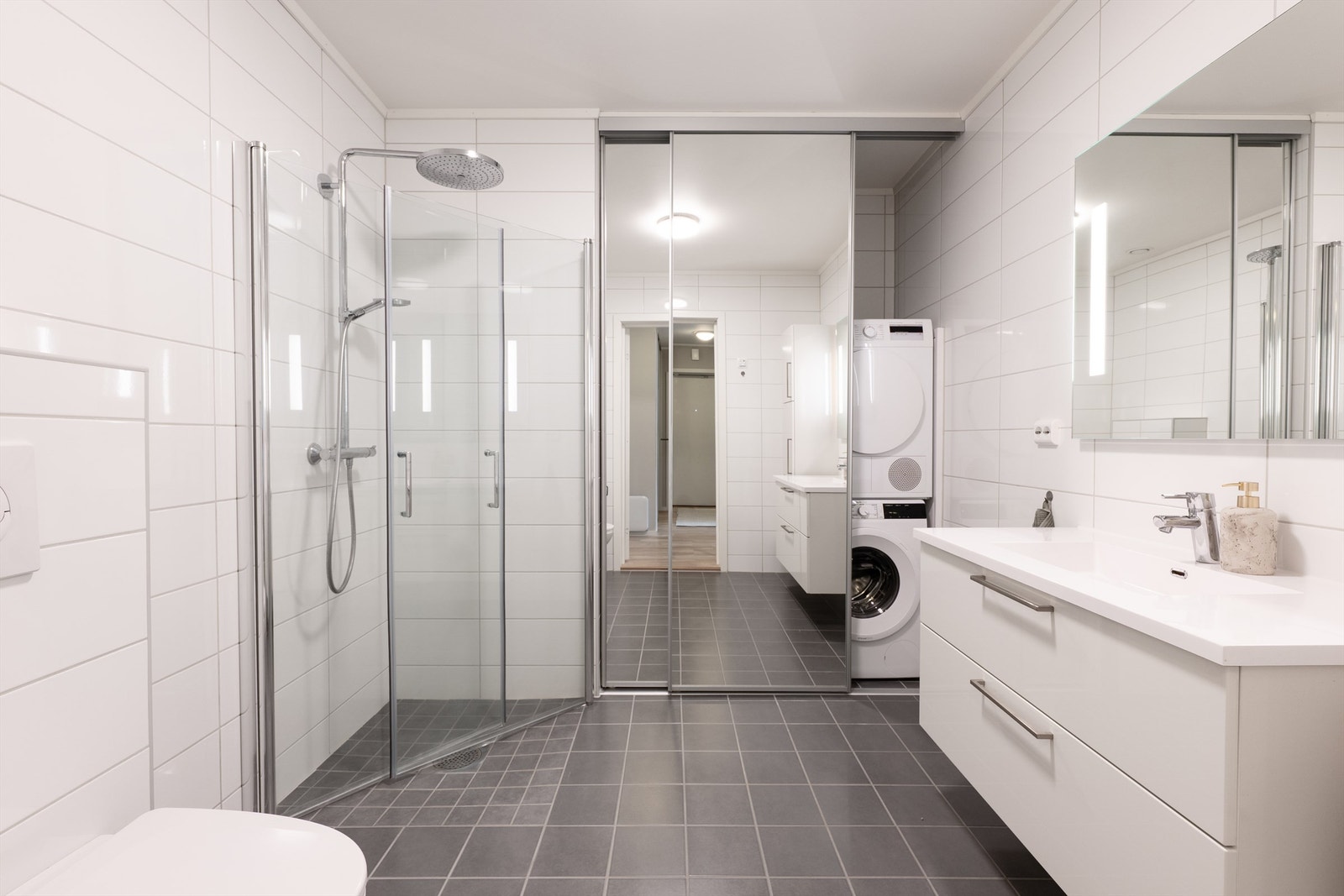 Badet har luftig følelse med mørke fliser, elegant glassdusj og store speilflater som forsterker lys og rom. Praktisk plassering av vaskemaskin/tørketrommel. Galleribilde