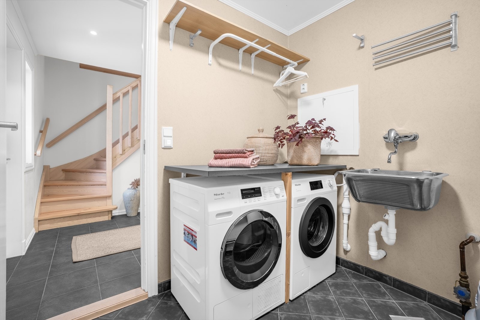 Praktisk vaskerom med fliser på betonggulv, tapet på veggene og slette overflater i himlingen. Det er opplegg for vaskemaskin og plass til tørketrommel. Galleribilde