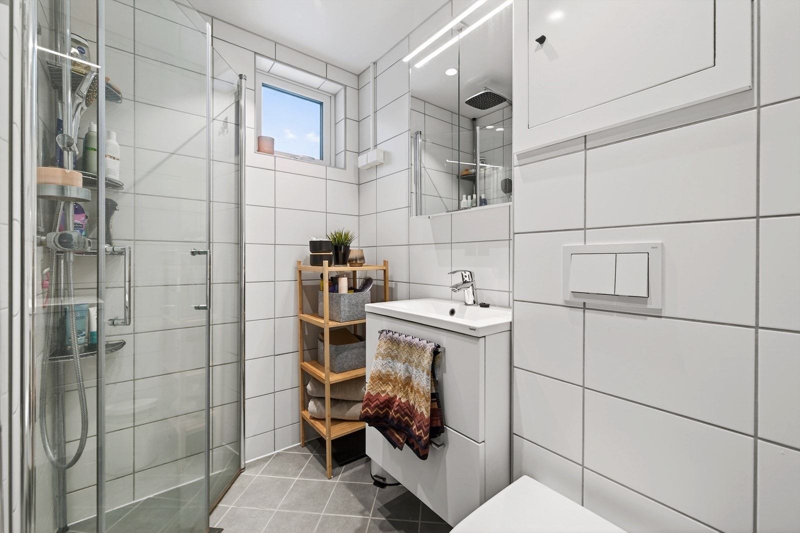 Baderomsinnredning med rom for oppbevaring i skuffeseksjoner og speil med belysning. Videre er badet utstyrt med toalett, dusjhjørne med innfellbare dusjvegger. Galleribilde