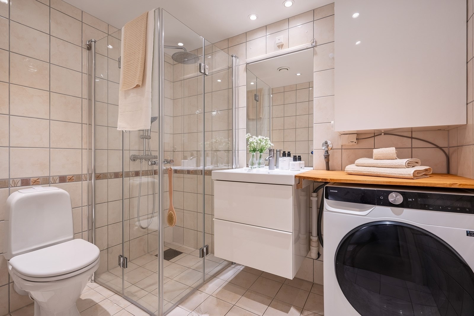 Lyst flislagt bad med varmekabler i gulv. Badet er innredet med servantskap, toalett, skap og hjørnedusj. Det er opplegg for vaskemaskin i hjørnet. Galleribilde