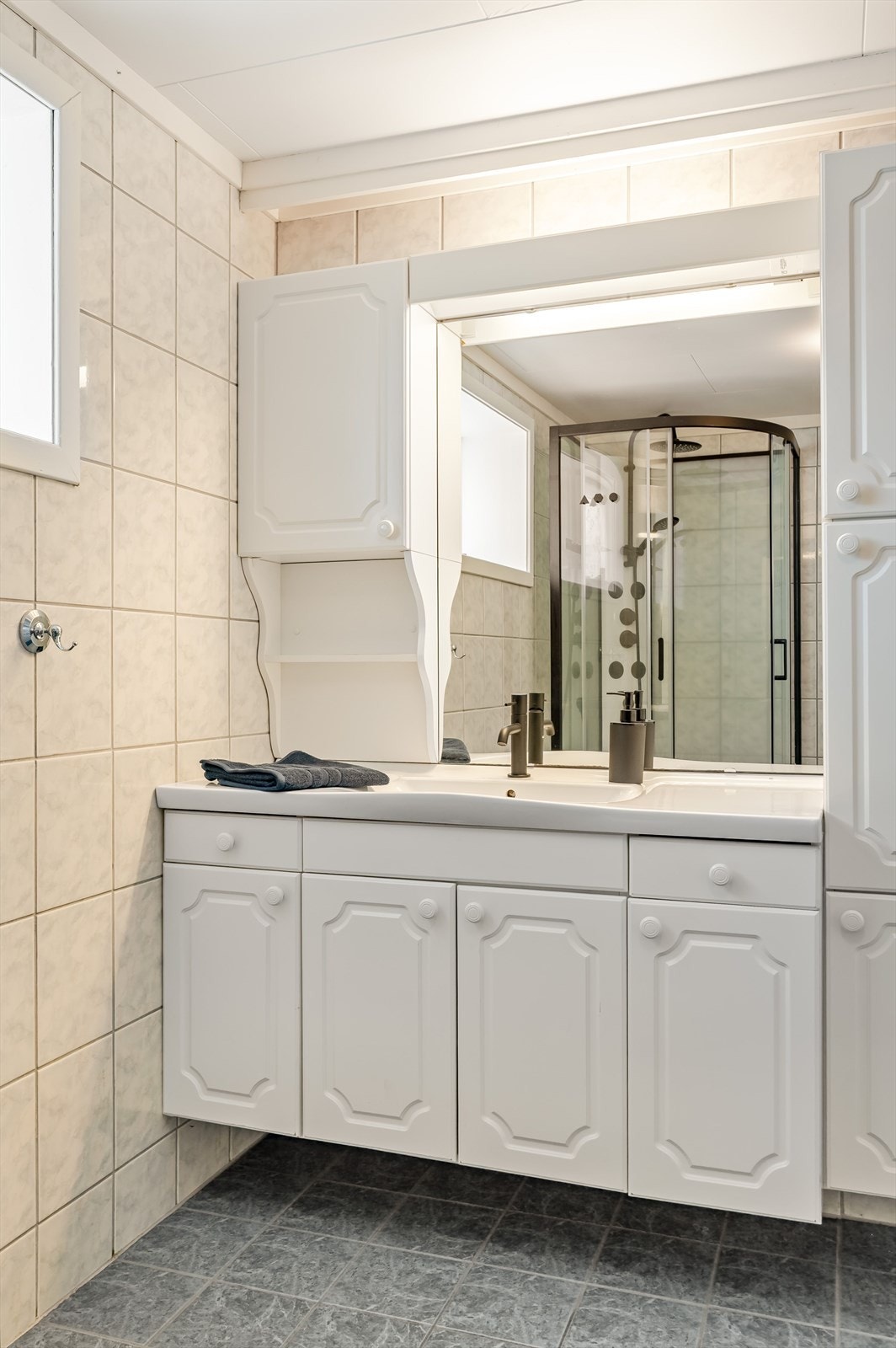 Lys baderomsinnredning med profilerte fronter og heldekkende servant med sort armatur, speil med belysning og vegghengt skap over. Baderommet er også utstyrt med dusjkabinett med både hånd- og regnfallsdusj, samt gulvstående toalett. Galleribilde