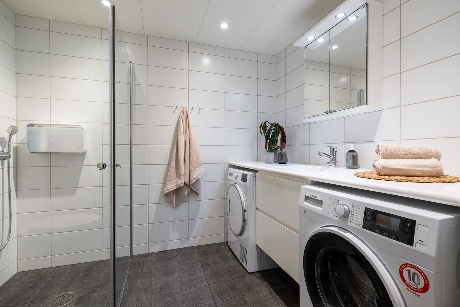 Lyst flislagt bad med vannbåren varme i gulv og downlights i himling. Det er opplegg for vaskemaskin og plass til tørketrommel under benk. Galleribilde