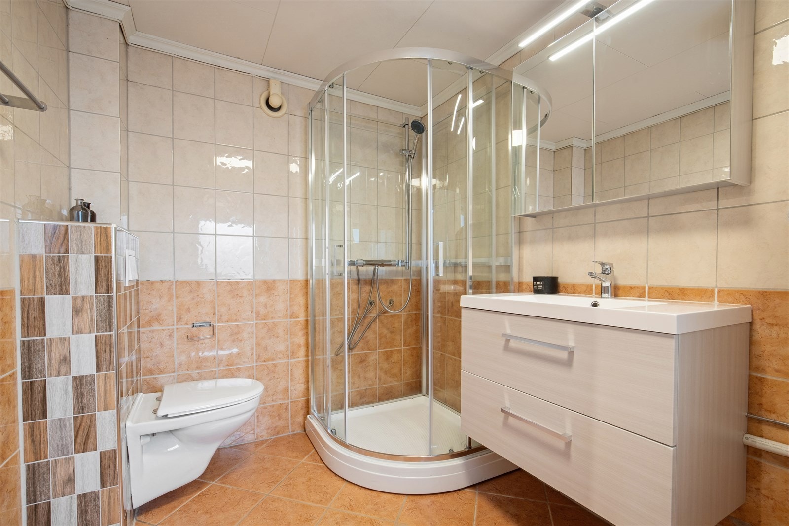 Badet er fra 90-tallet og er innredet med en kombinasjon av hvite og brune fliser på gulv og vegger. I gulvet har man også varmekabler for ekstra komfort. Galleribilde