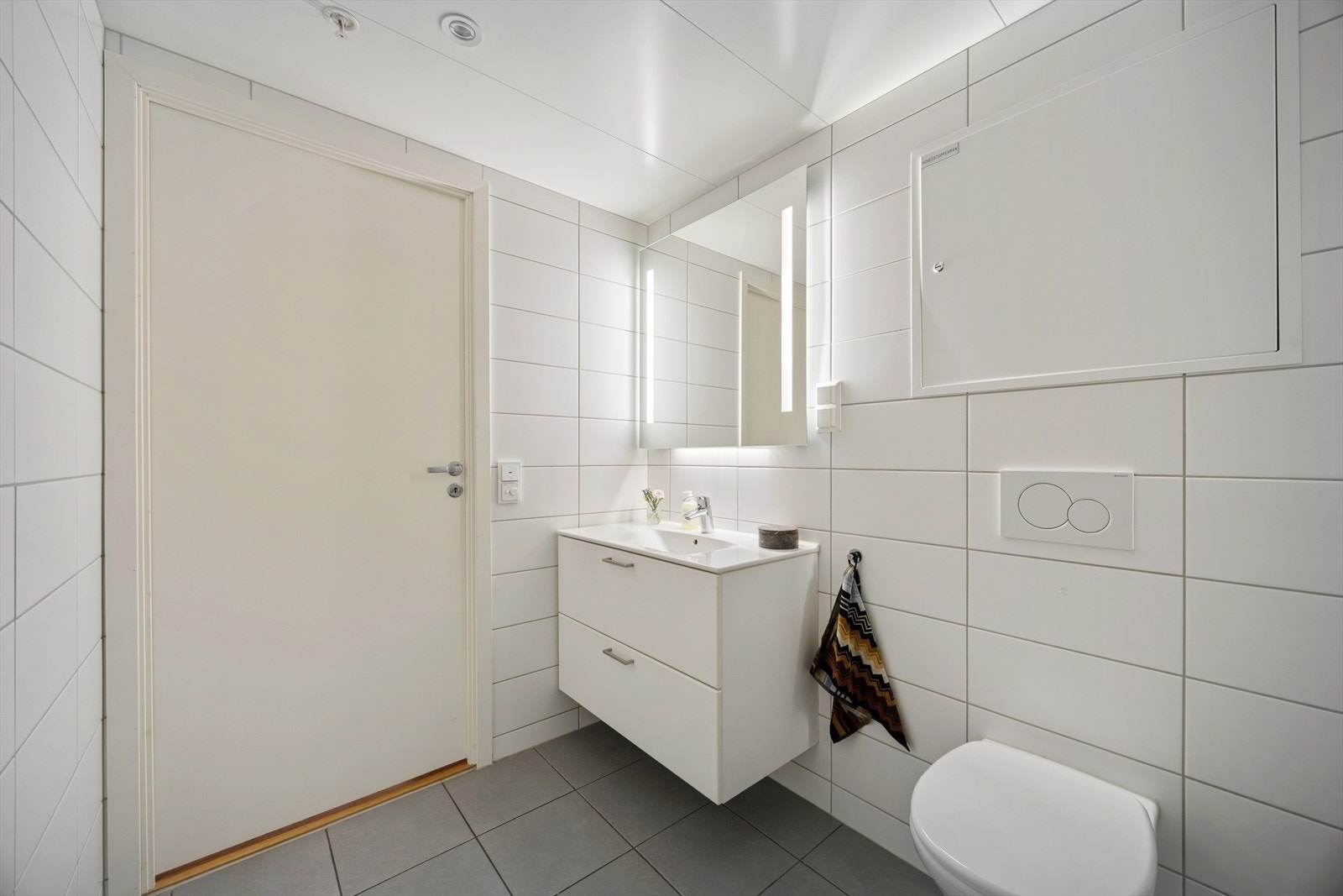 Badet består av vegghengt toalett, dusjhjørne med dør og en servantinnredning med heldekkende servant og skuffer. Over servanten er det speil med integrert belysning. Galleribilde