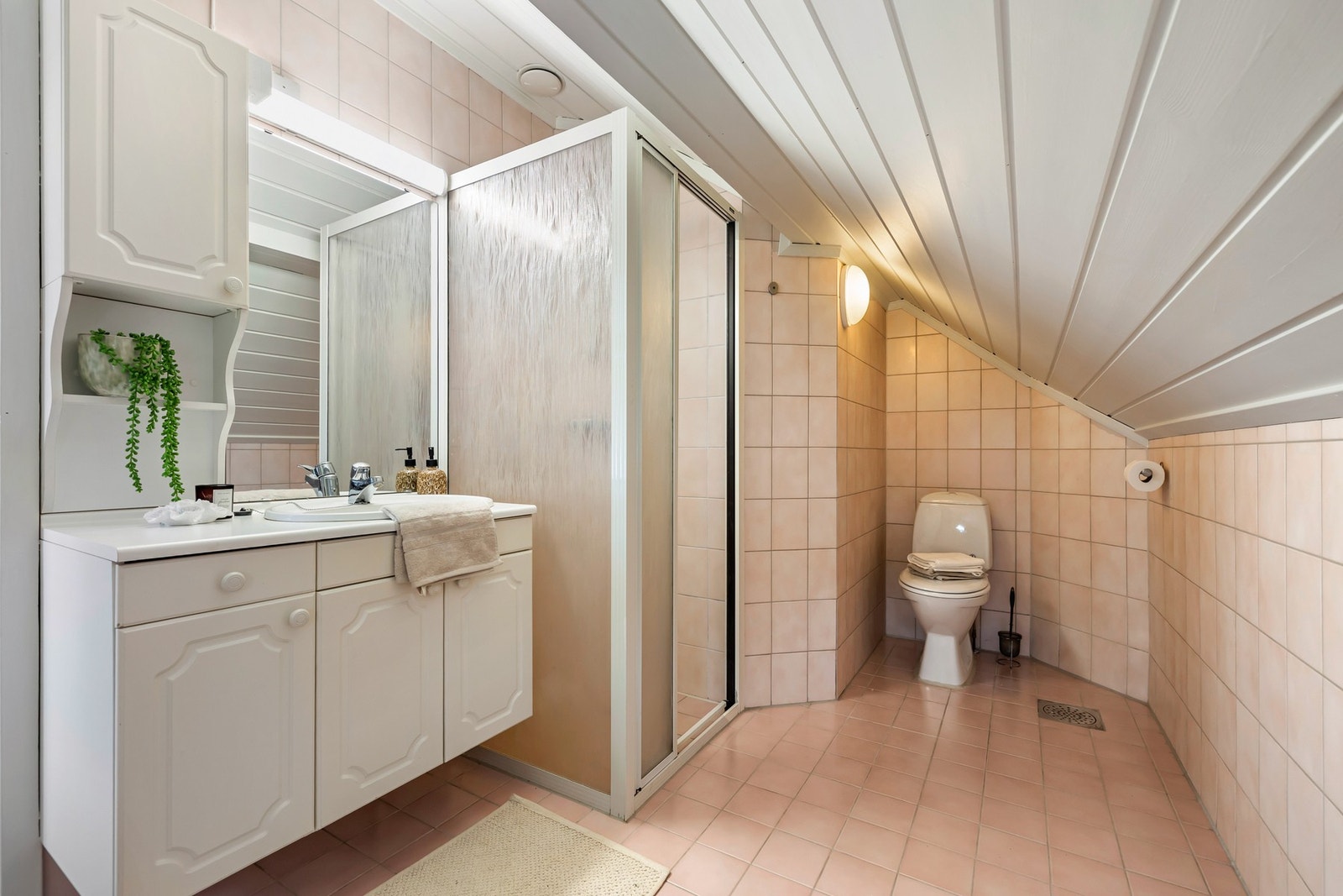 Rommet er flislagt på gulv og vegger i lyse toner. Badet er innredet med dusjkabinett, toalett og servant med underskap og speilskap over. Galleribilde