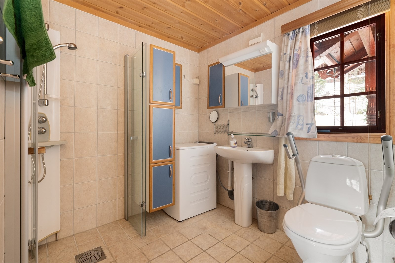 Badet inneholder servant, overskap, høyskap, overskap med speil og lys, dusjhjørne med glassdører, gulvmontert toalett og opplegg til vaskemaskin Galleribilde