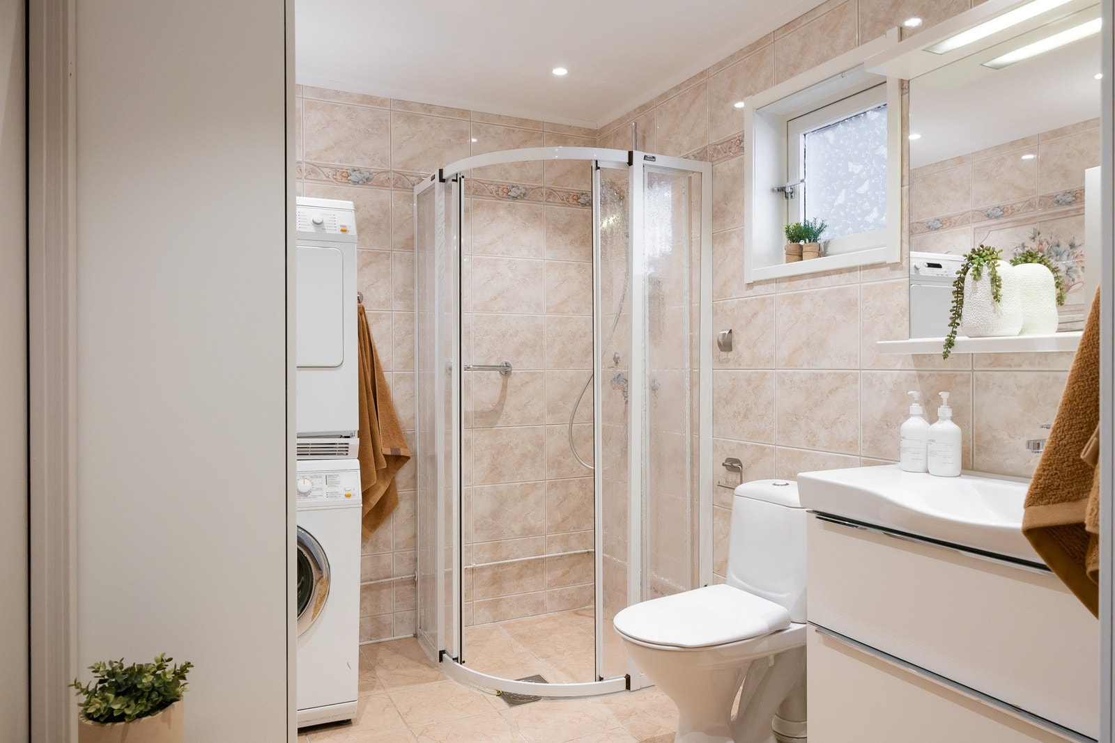 Flislagt baderom med servantmøbler med skuffer, veggspeil med belysning, dusjhjørne og gulvstående toalett. Opplegg for vaskemaskin. Galleribilde