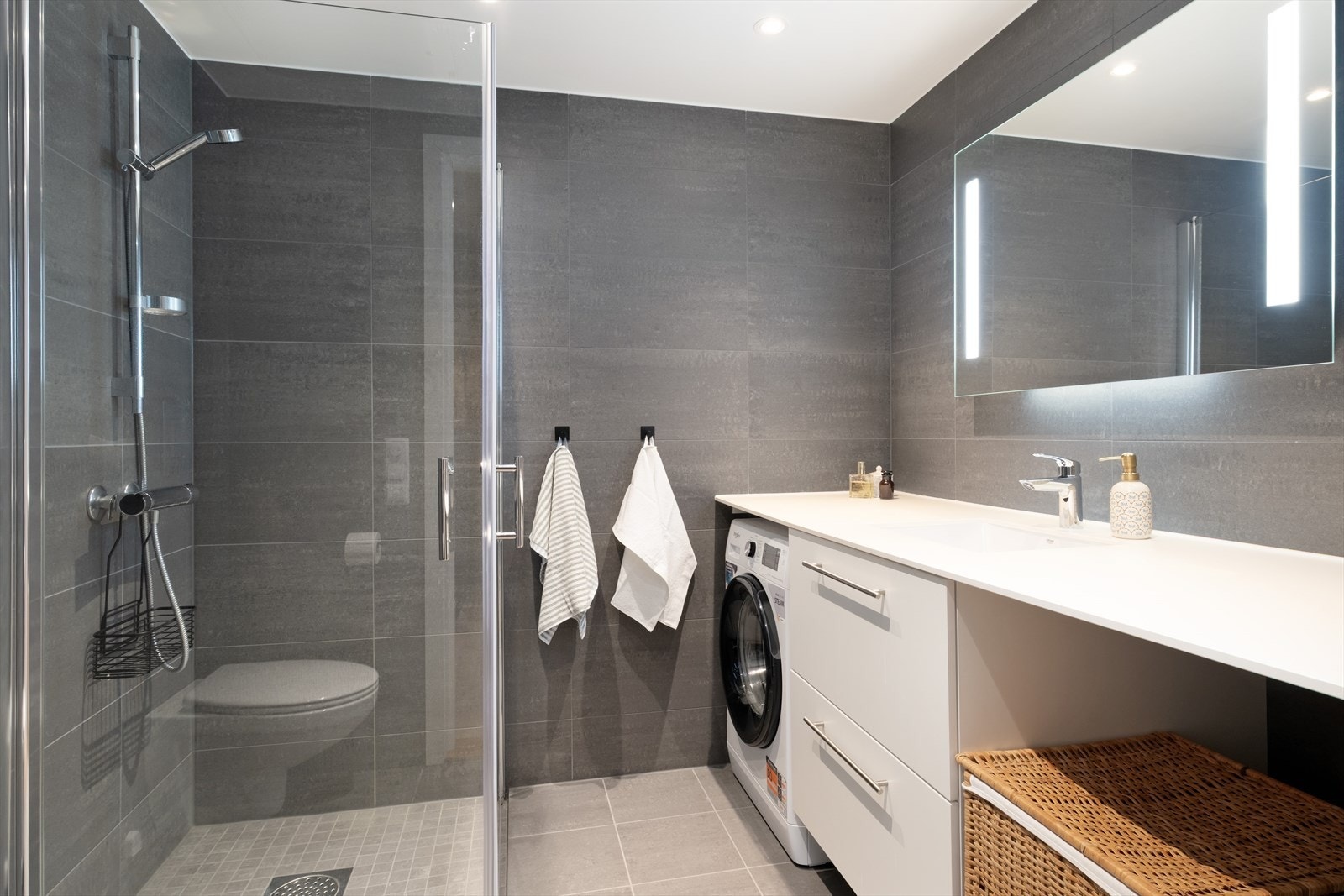 Bad/vaskerom med flisar på golv og veggar. Her har ein vegghengt toalett, dusjnisje, servantskap og opplegg for vaskemaskin under benkeplate. Galleribilde