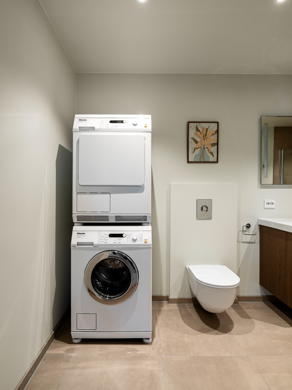 Store fliser på gulv og dusjsone i lun farge. Elegant dusjløsning med nedsenket gulv, designsluk og glassvegger som kan foldes mot vegg. Veggmontert toalett og servant i skuffeseksjon med høyskap. Innfelt led-belysning og opplegg til vaskemaskin. Galleribilde