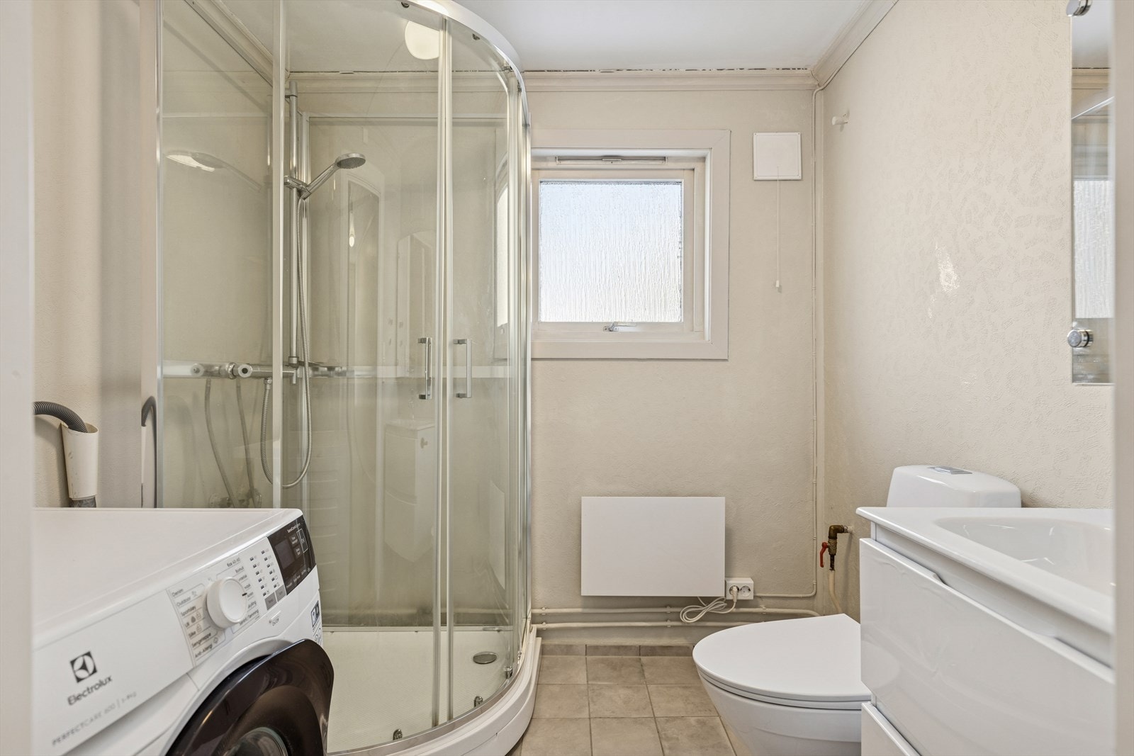 Enkelt baderom, utstyrt med dusjkabinett, heldekkende servant, gulvstående toalett, opplegg for vaskemaskin og panelovn. Galleribilde