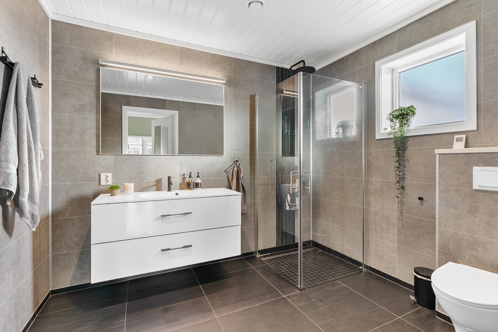 Pent og romslig bad innredet med vegghengt toalett, dusj med glassdører og regndusj, samt baderomsinnredning med skuffer og heldekkende servant. Galleribilde