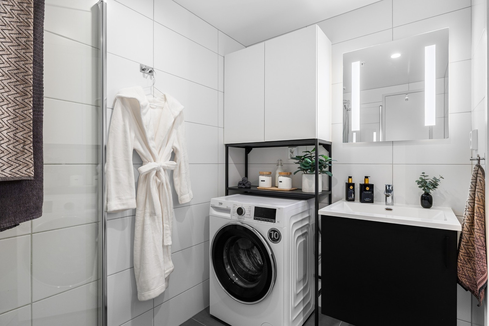 Innredet med dusjdører i glass, dusjbatteri og garnityr montert på vegg, veggmontert WC, Kran og avløp for vaskemaskin, samt rørskap over toalettet. Galleribilde