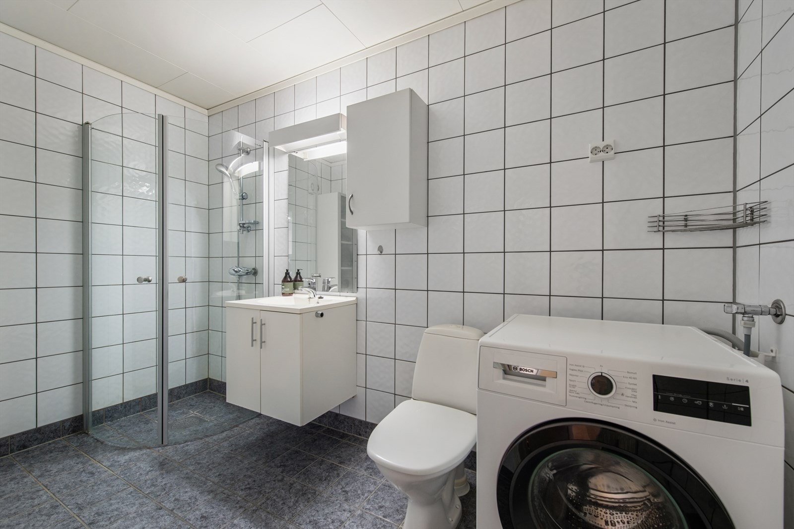 Badet er utstyrt med toalett, dusjhjørne med glassdører, baderomsinnredning med servant, samt opplegg for vaskemaskin. Galleribilde