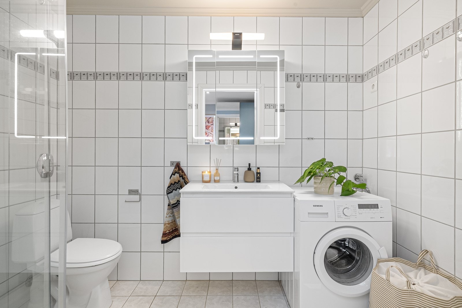 Lyst, flislagt bad med standard fra byggeår. Nyere, pen servantinnredning med skuffer og heldekkende servant, samt speilskap med belysning. Galleribilde