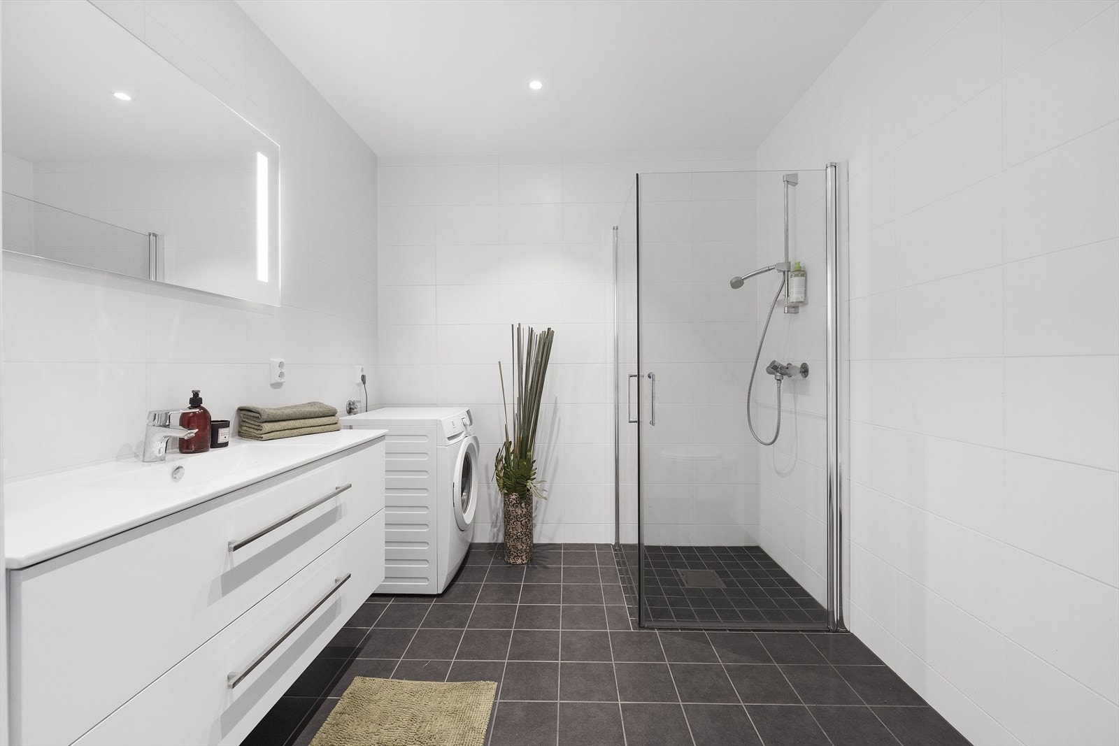 Pent flislagt baderom med varmekabler i gulv. Rommet er utstyrt med servant over skuffeinnredning, speil med belysning, toalett, dusjhjørne med innfellbare glassvegger, samt opplegg til vaskemaskin og tørketrommel Galleribilde