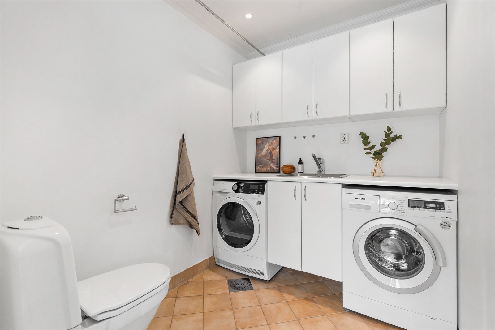 Vaskerom med flislagt gulv med varme. Nedsenket himling med downlights. Vegghengt benkeskap. Nedfelt utslagsvask med ett-greps armatur. Opplegg for vaskemaskin og tørketrommel. Gulvstående toalett. Galleribilde