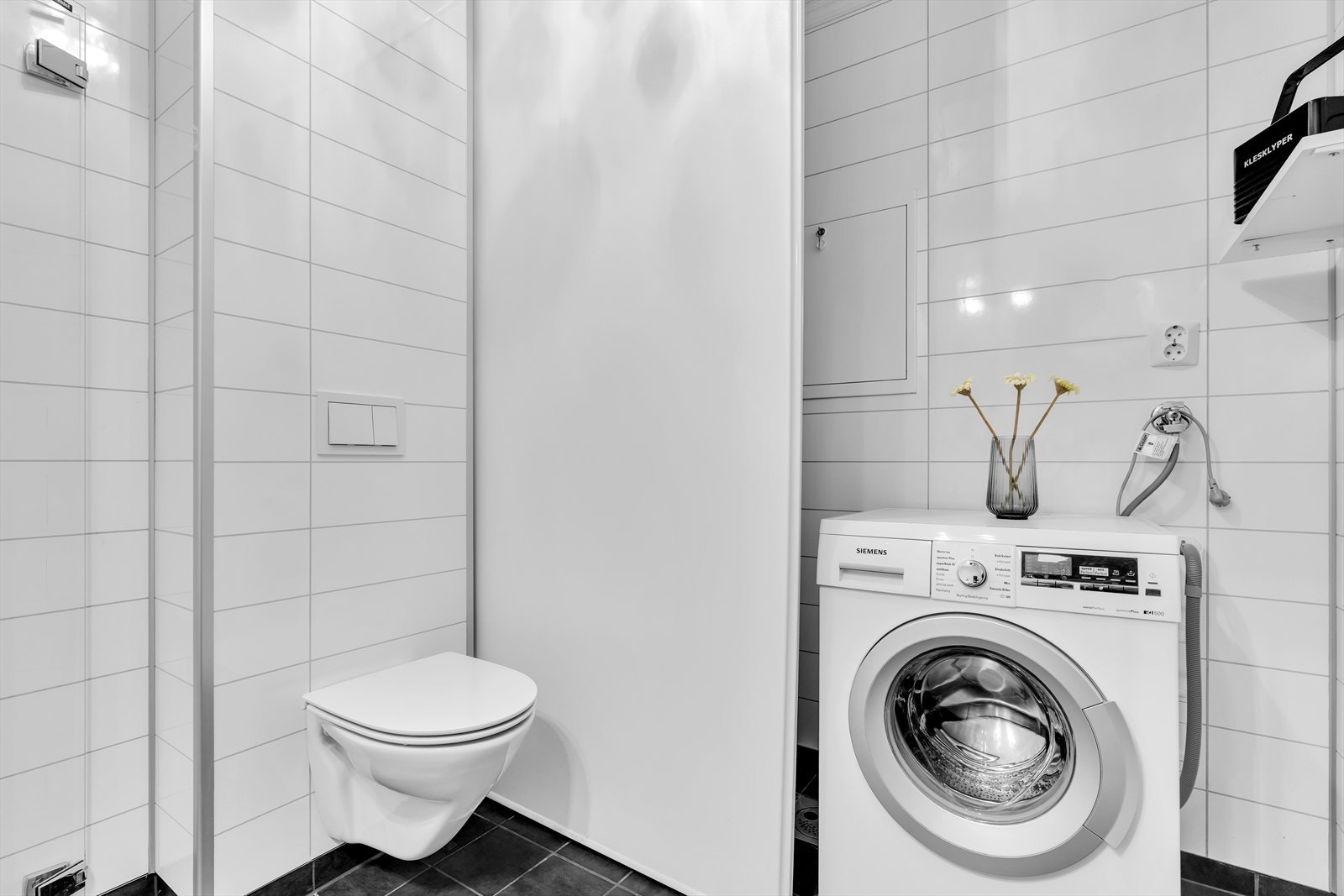 Badet inkluderer en praktisk innredning med opplegg for vaskemaskin, vegghengt toalett og dusjløsning med dusjdør og garnityr. Galleribilde