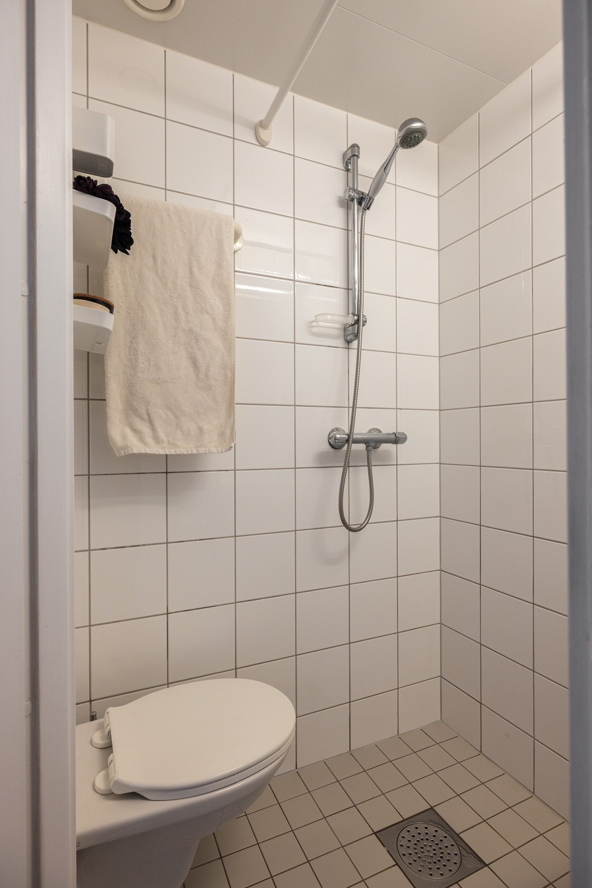 Lyse fliser på både gulv og vegger. Vegghengt servant, speil med glasshylle og belysning over, dusjhjørne med forheng og veggmontert toalett. Galleribilde